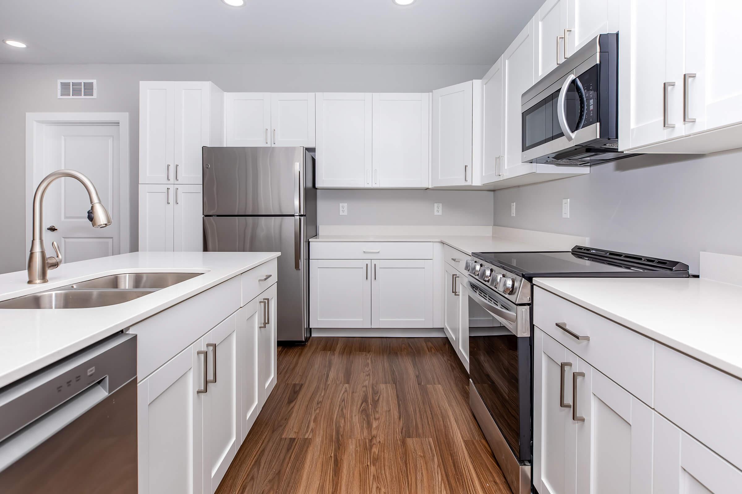 A modern kitchen featuring white cabinetry, stainless steel appliances, including a refrigerator, stove, and microwave, with a farmhouse-style sink. The countertops are a light color, and the floor is made of hardwood, creating a contemporary and clean aesthetic.