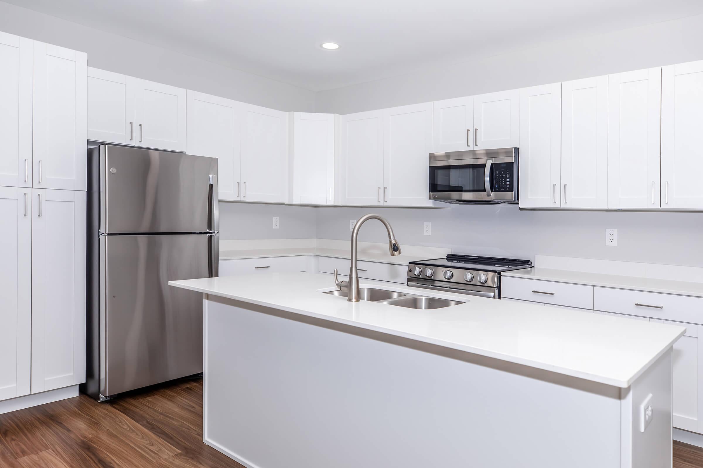 Modern kitchen featuring white cabinetry, stainless steel refrigerator, microwave, and stove. The island has a double sink and a sleek countertop, with light wood flooring and bright overhead lighting. The design emphasizes a clean and contemporary aesthetic.