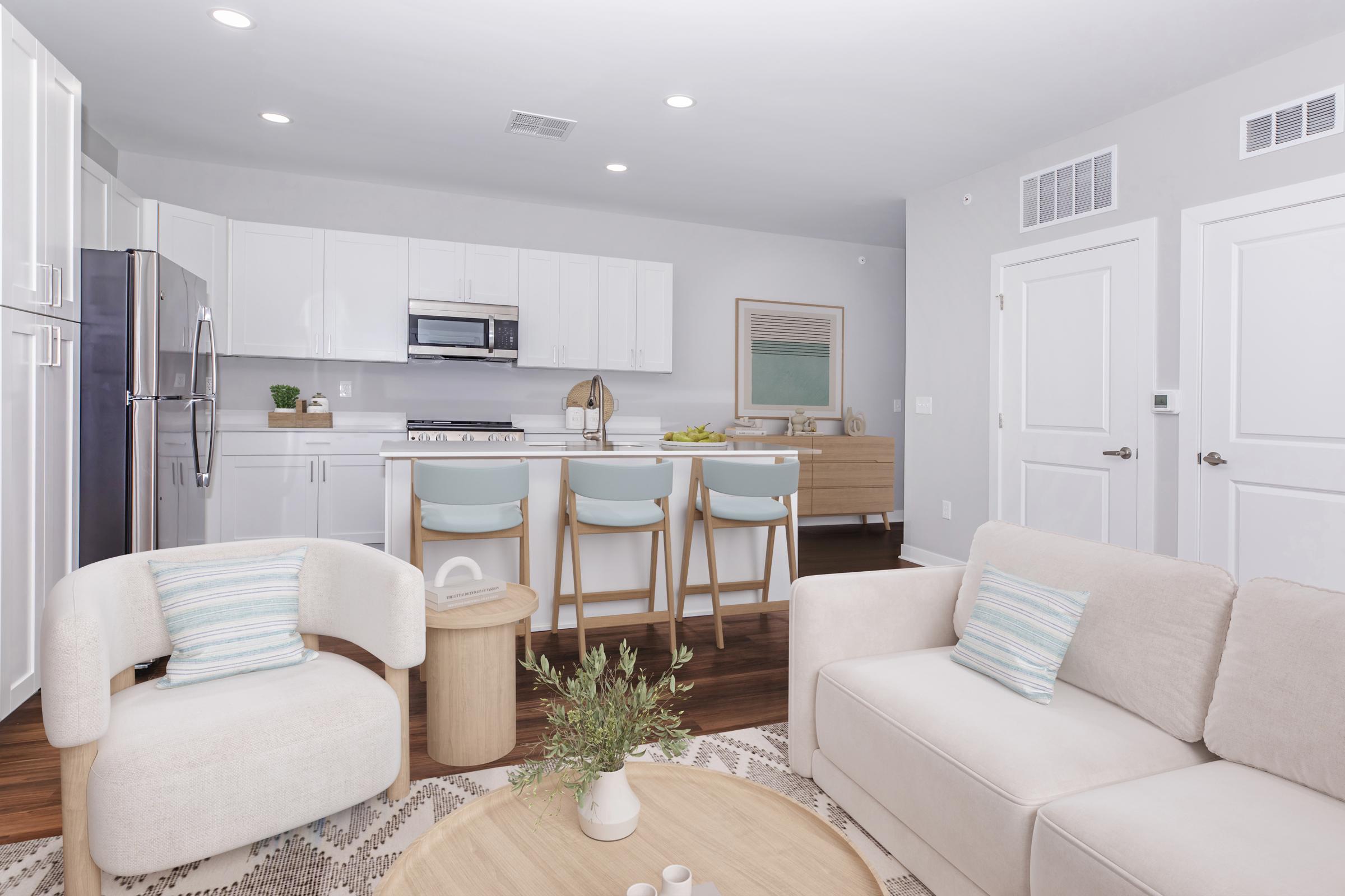 Modern kitchen and living area featuring sleek white cabinetry, stainless steel appliances, and a breakfast bar with light blue stools. The living space includes a light-colored sofa and armchair, a round wooden coffee table, and decorative elements like a plant and a patterned rug. Bright, airy atmosphere.