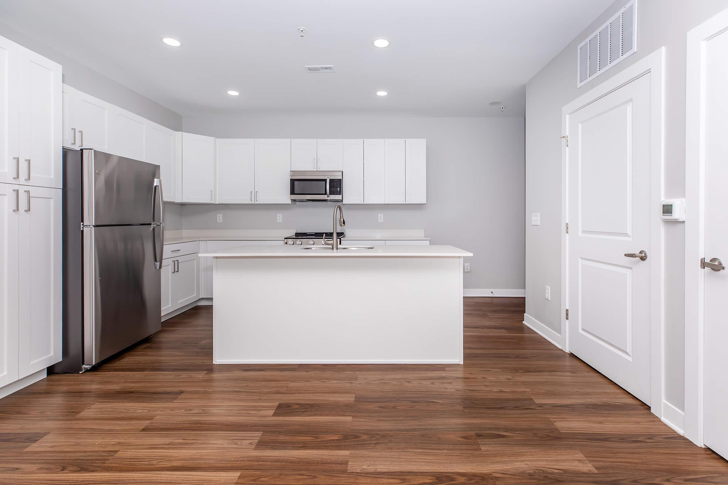Modern kitchen with stainless steel appliances, including a refrigerator and microwave. Features white cabinetry, a large island with a sink, and hardwood flooring. The walls are painted light gray, and the space is well-lit with recessed lighting.