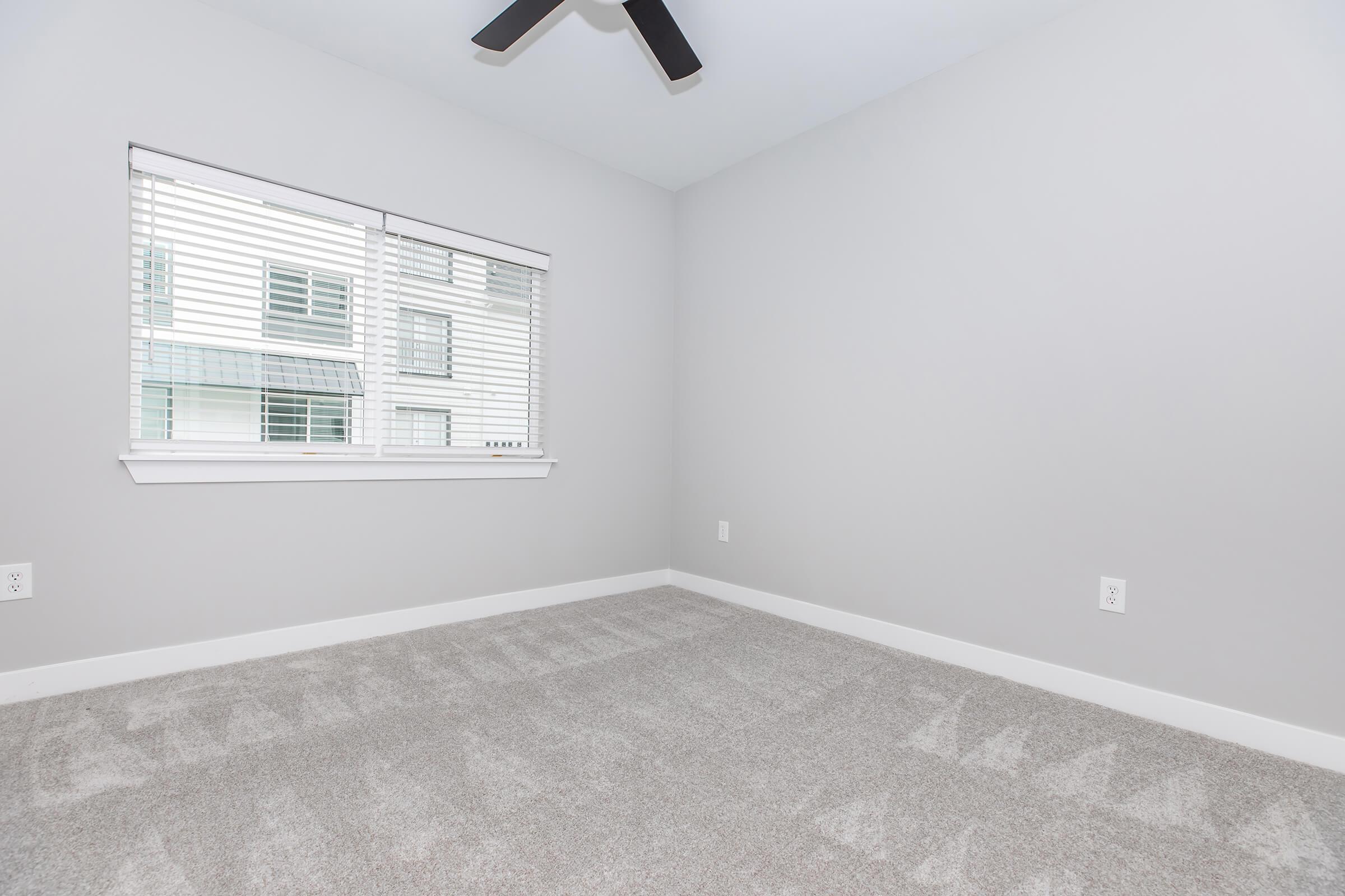 A spacious, empty room with light gray walls and carpeted floor. A window with white blinds allows natural light to enter, providing a view of an exterior building. The ceiling features a black ceiling fan, adding a modern touch to the minimalist design.