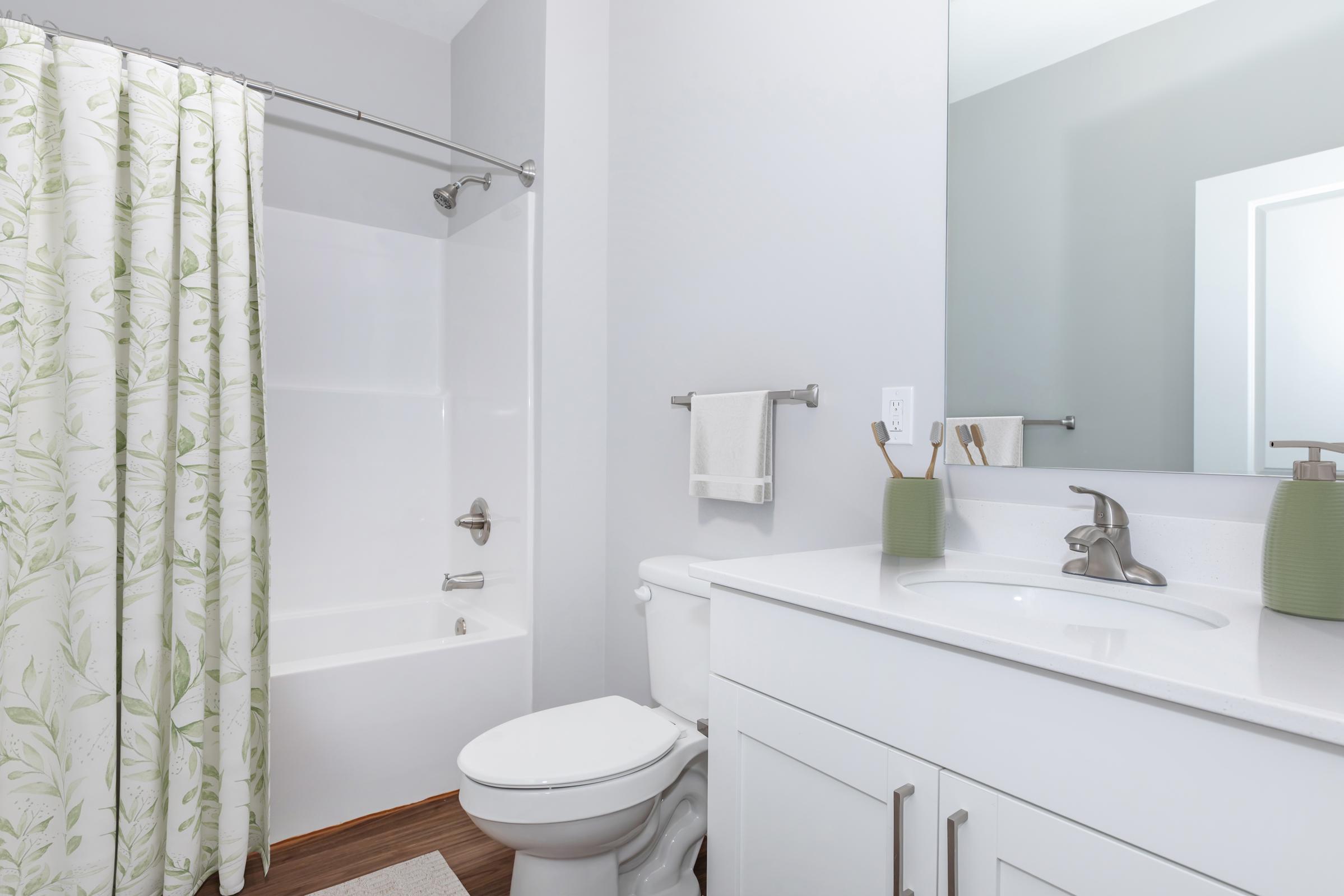 A modern bathroom featuring a white bathtub and shower with a patterned green and white curtain, a sleek white vanity with a sink, and a large mirror. The decor includes green accents, a towel rack, and a minimalist design, creating a clean and fresh aesthetic.