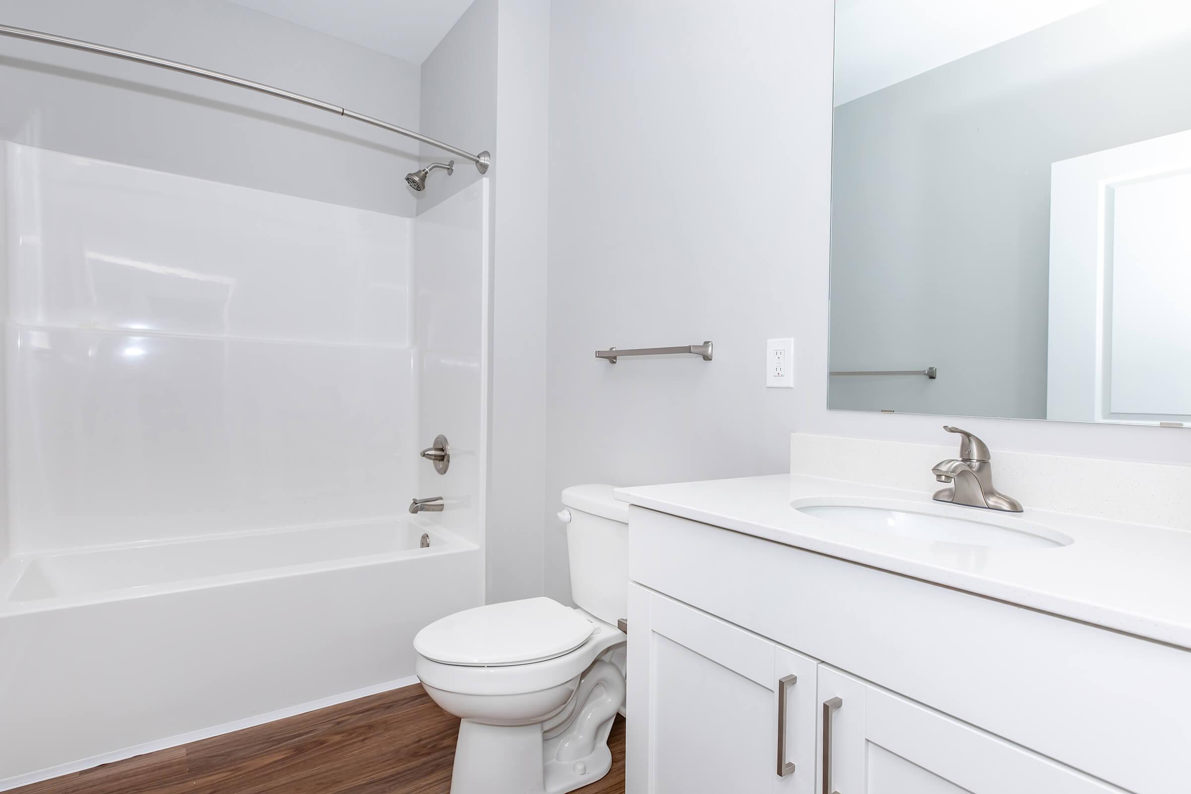 A clean and modern bathroom featuring a white bathtub with a shower, a toilet, and a vanity with a sink. The walls are painted light gray, and the flooring is brown. There is a large mirror above the sink, and a towel bar is mounted on the wall.