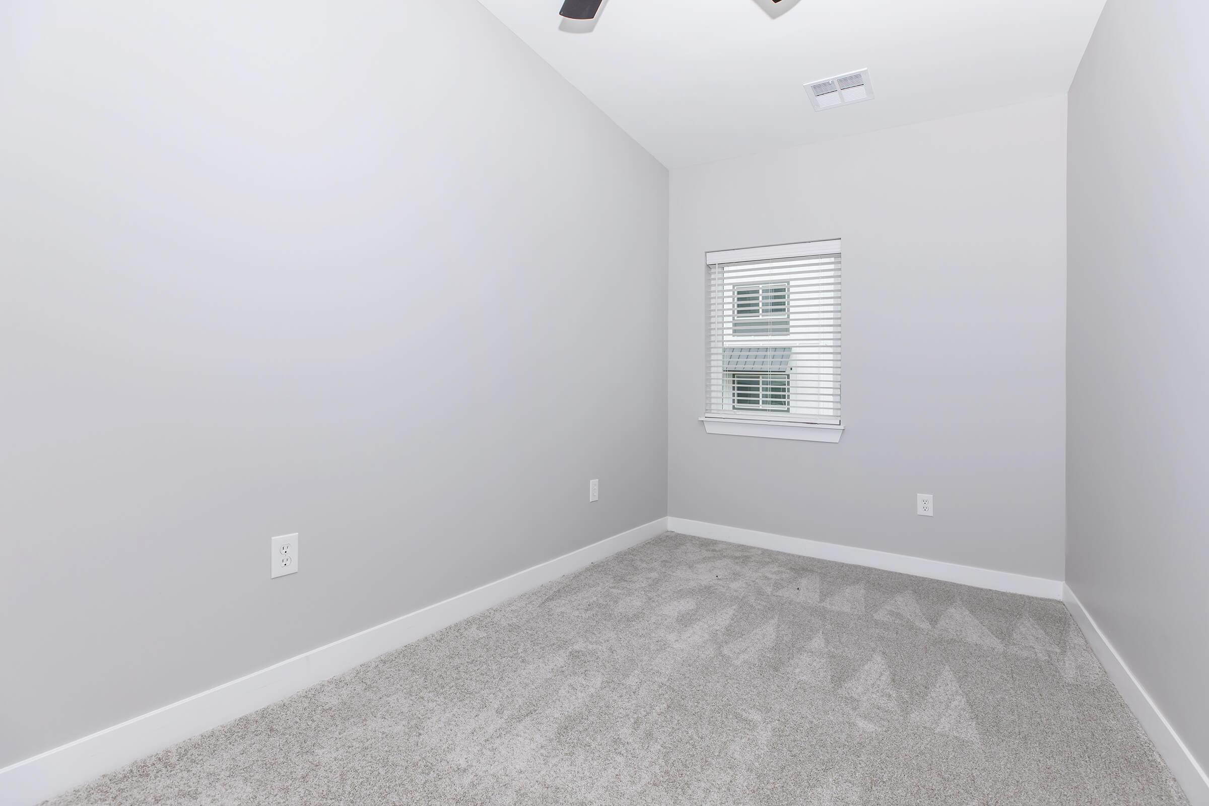 A small, empty room with light gray walls and carpet. There is a window with blinds on one wall, letting in natural light. The ceiling has a fan, and the overall atmosphere is minimalistic and clean, providing a blank canvas for potential decoration or use.