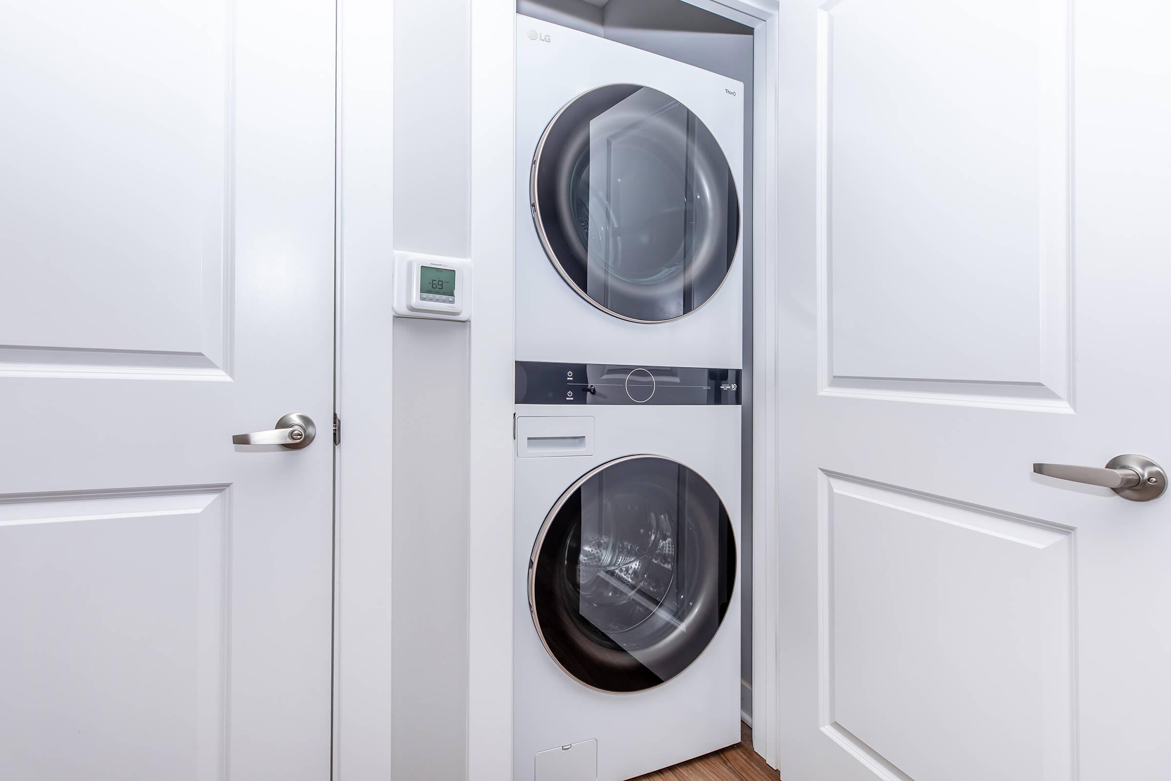 A stacked washer and dryer unit in a laundry room, positioned between two closed white doors. A wall-mounted thermostat is visible on the left side. The overall space is modern and well-lit, featuring light-colored walls and flooring.
