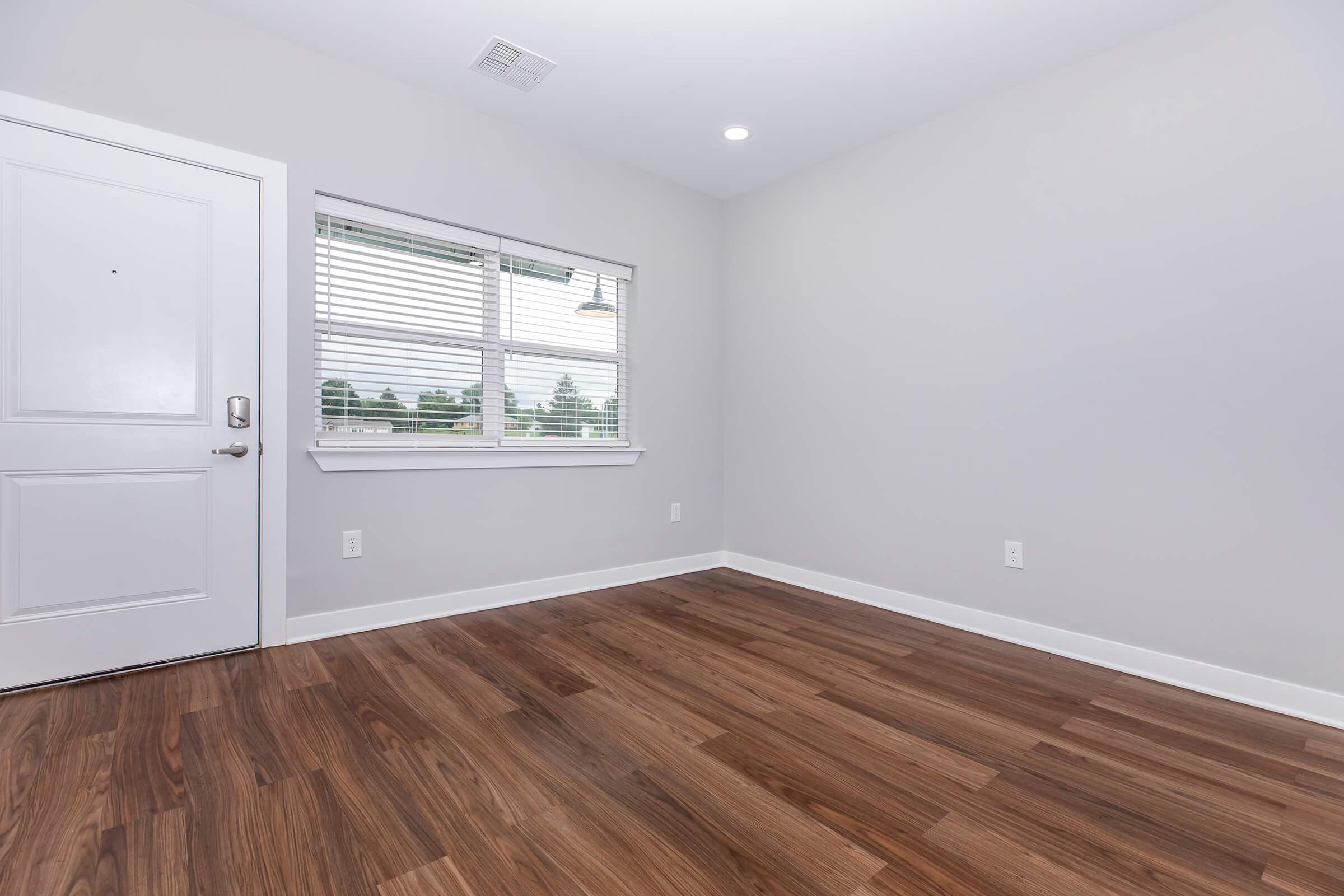 A bright, empty room featuring a light gray wall color, a door with a small window, and a single window with blinds. The flooring is made of light brown hardwood, creating a warm atmosphere. Natural light enters through the window, illuminating the space.