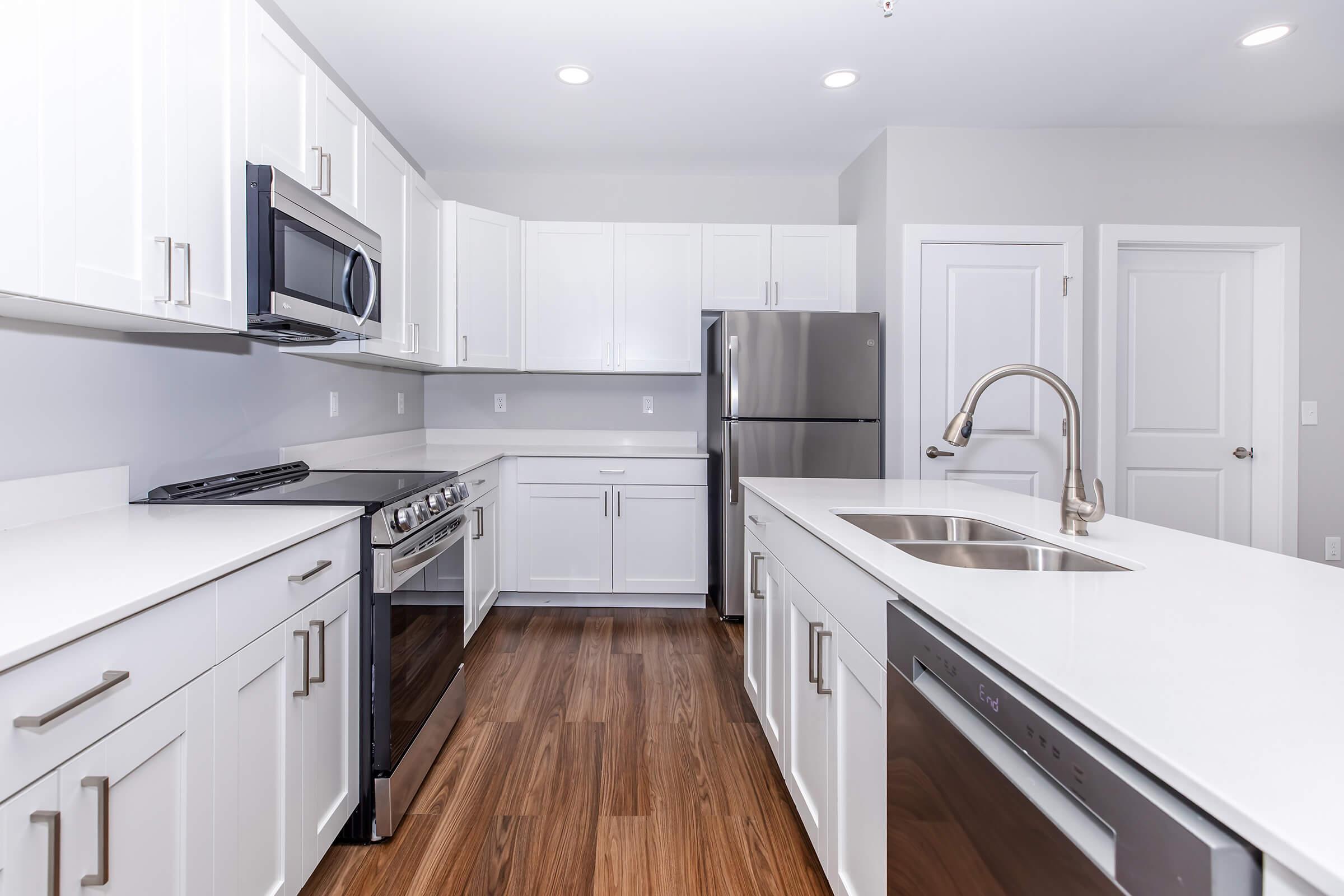 Modern kitchen featuring white cabinetry, stainless steel appliances including a refrigerator and microwave, a sleek gas stove, and a stainless steel sink. The countertops are white, complemented by wooden flooring. The space is bright with recessed lighting and a minimalist design.
