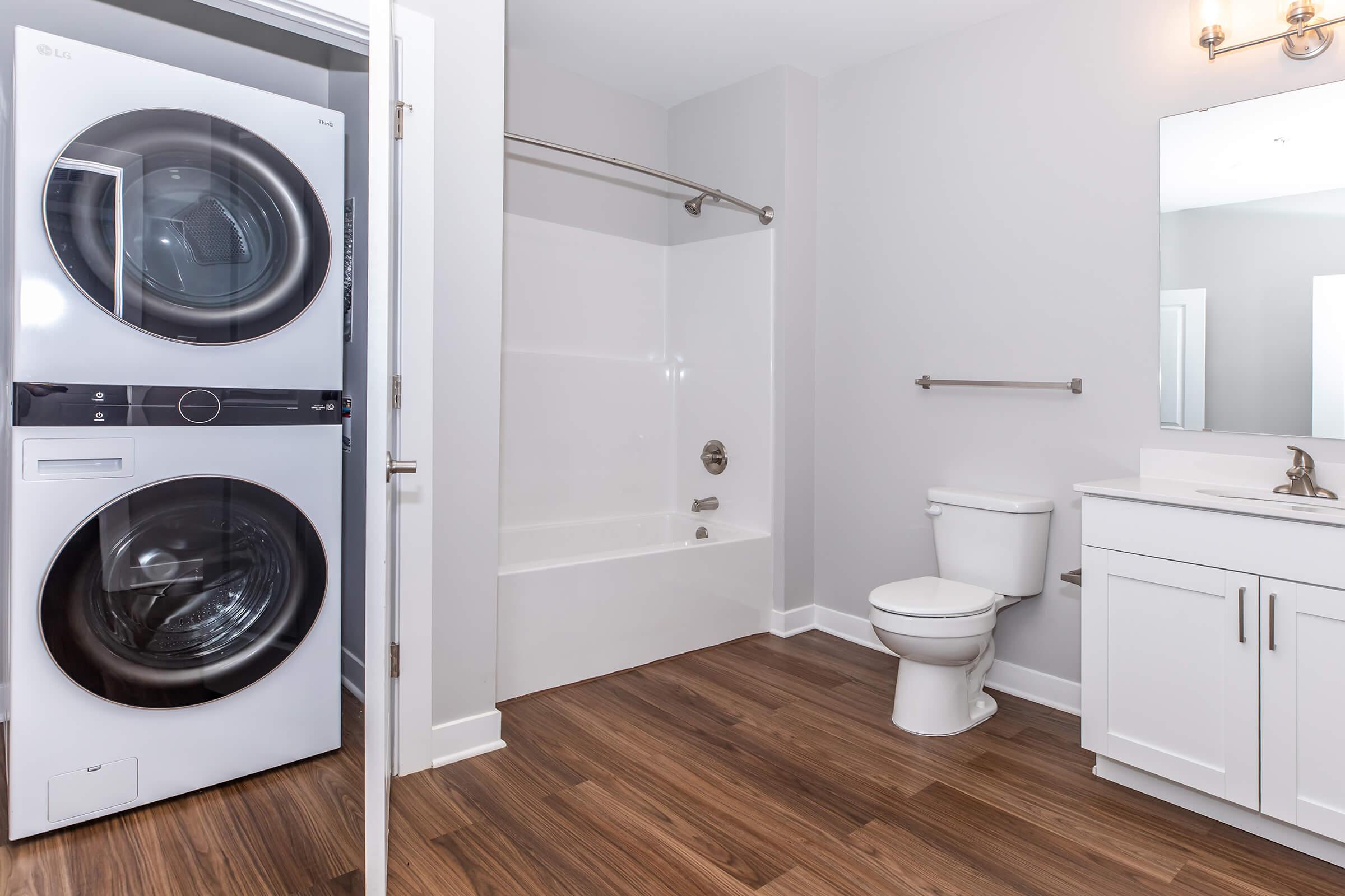 A modern bathroom featuring a white bathtub and shower combo, a toilet, and a vanity with a sink and mirror. To the left, there is a stacked washer and dryer unit. The flooring is a warm wooden finish, and the walls are painted a light gray, creating a contemporary and clean look.
