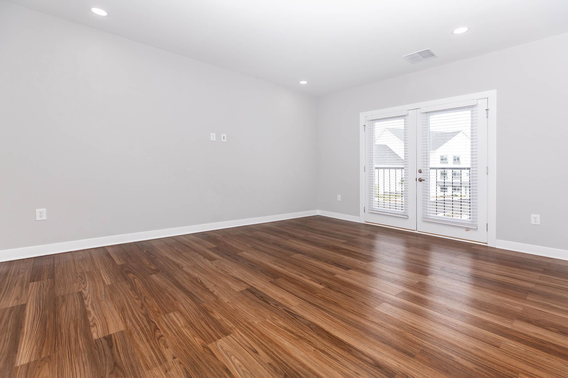 A spacious, empty room with light gray walls and hardwood flooring. There are two sets of double doors leading to a small balcony outside. The room is brightly lit with recessed ceiling lights, creating an airy atmosphere.