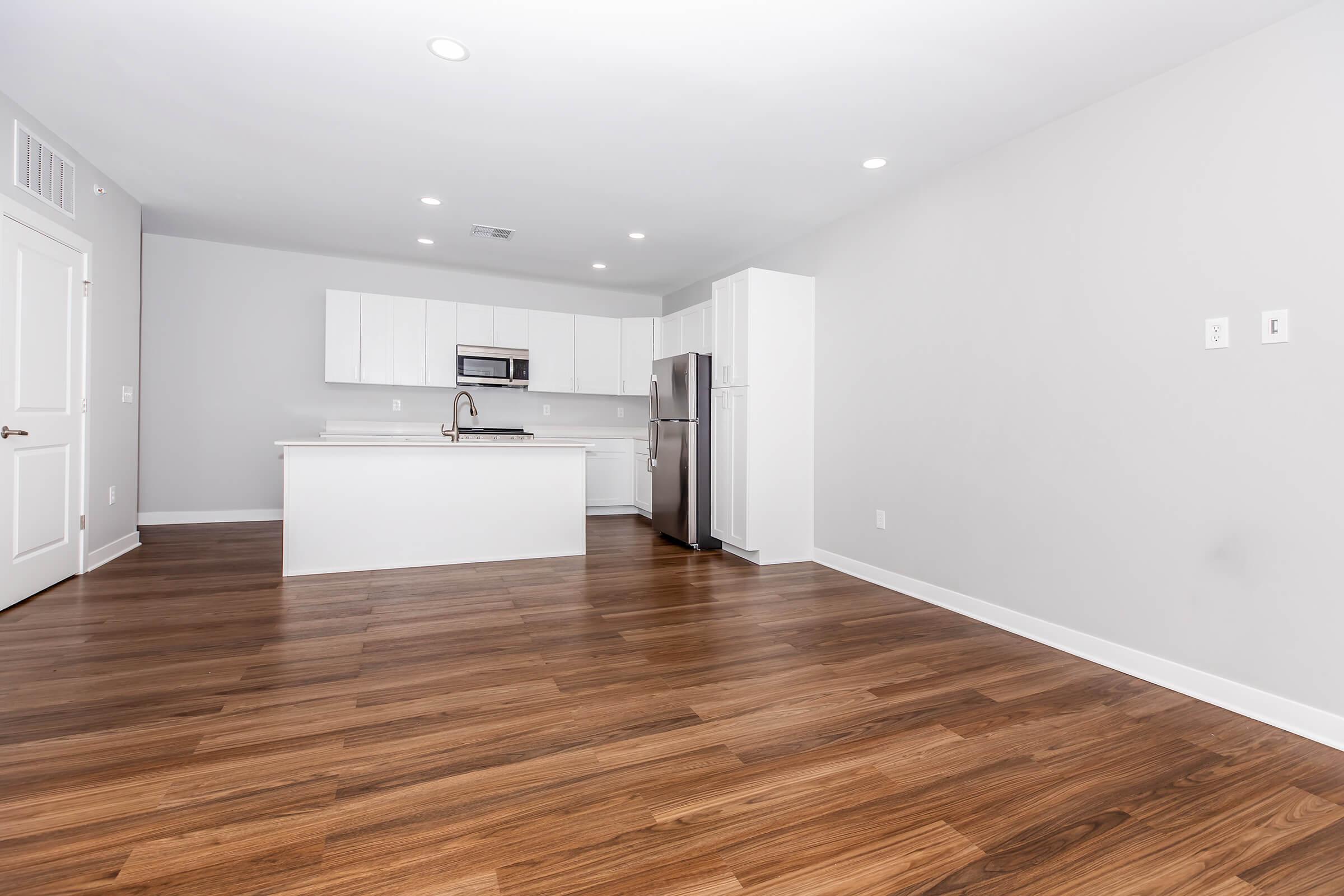 Spacious, modern kitchen and living area with light wood flooring, white cabinetry, and stainless steel appliances. The open layout features ample natural light, perfect for entertaining. Neutral-colored walls create a bright and inviting atmosphere.