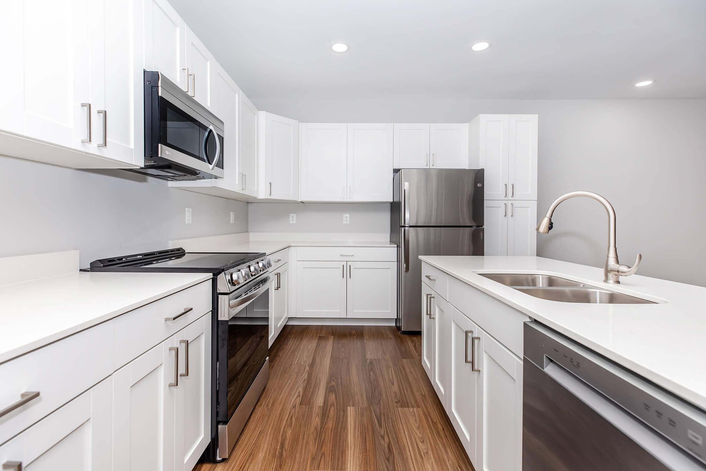 Modern kitchen featuring white cabinetry, stainless steel appliances, a gas range, a spacious fridge, and a double-basin sink. The countertops are a light color, and the flooring is hardwood, creating a bright and contemporary atmosphere. Ample storage space is available with organized cabinetry.