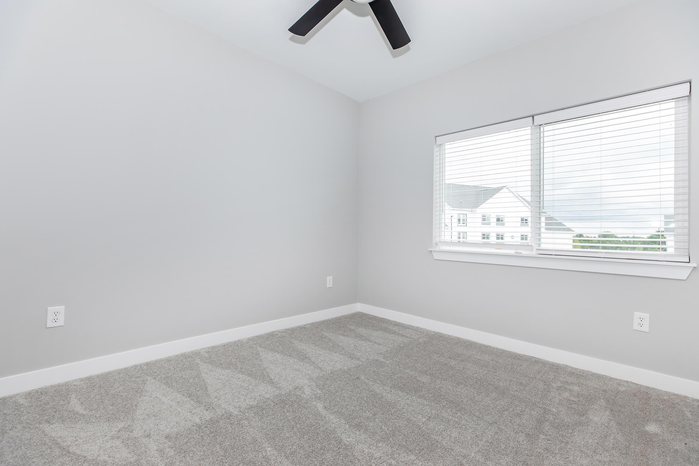 A simple, empty room with light gray walls and carpet. A ceiling fan is visible, and a window with blinds lets in natural light, revealing a view of an exterior building. The room is sparsely decorated, creating a clean and minimalist space.