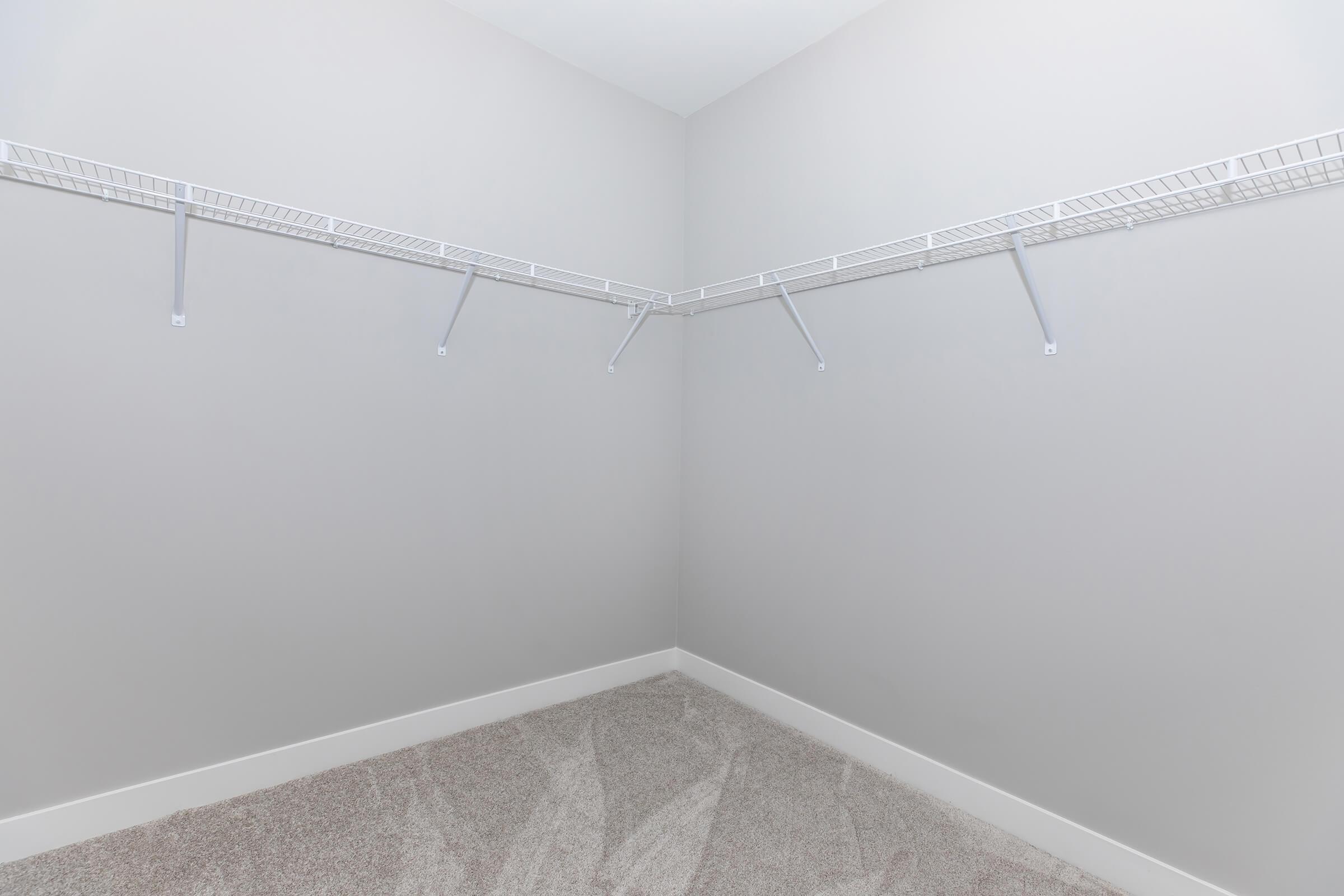 Empty walk-in closet with light gray walls and carpeted flooring. Two wire shelving units are mounted to the walls, providing storage options. The space is bright and clean, creating a minimalistic look.