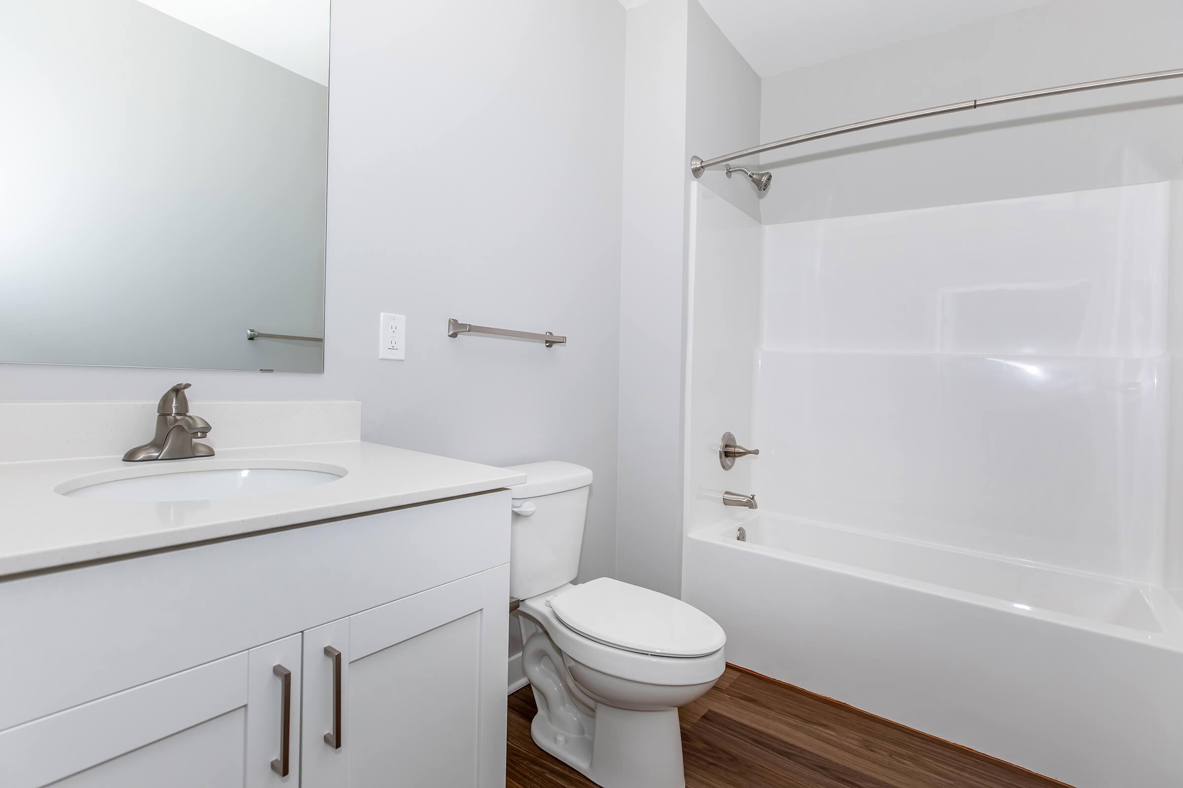 A modern bathroom featuring a white sink with a mirror above, a toilet, and a clean bathtub with a shower. The walls are painted light gray, and the floor has wooden accents.