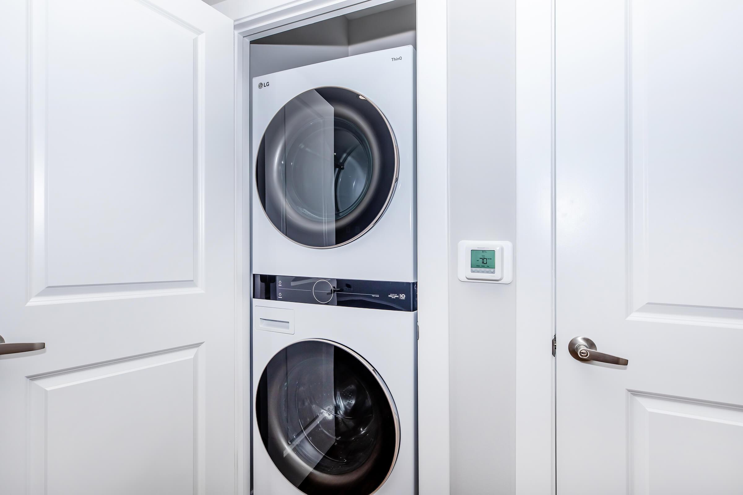 A modern stackable washer and dryer set in a narrow laundry closet with white doors. The appliances are sleek and designed for space efficiency, featuring a digital control panel visible on the front of the dryer. A thermostat is located on the wall beside the appliances.
