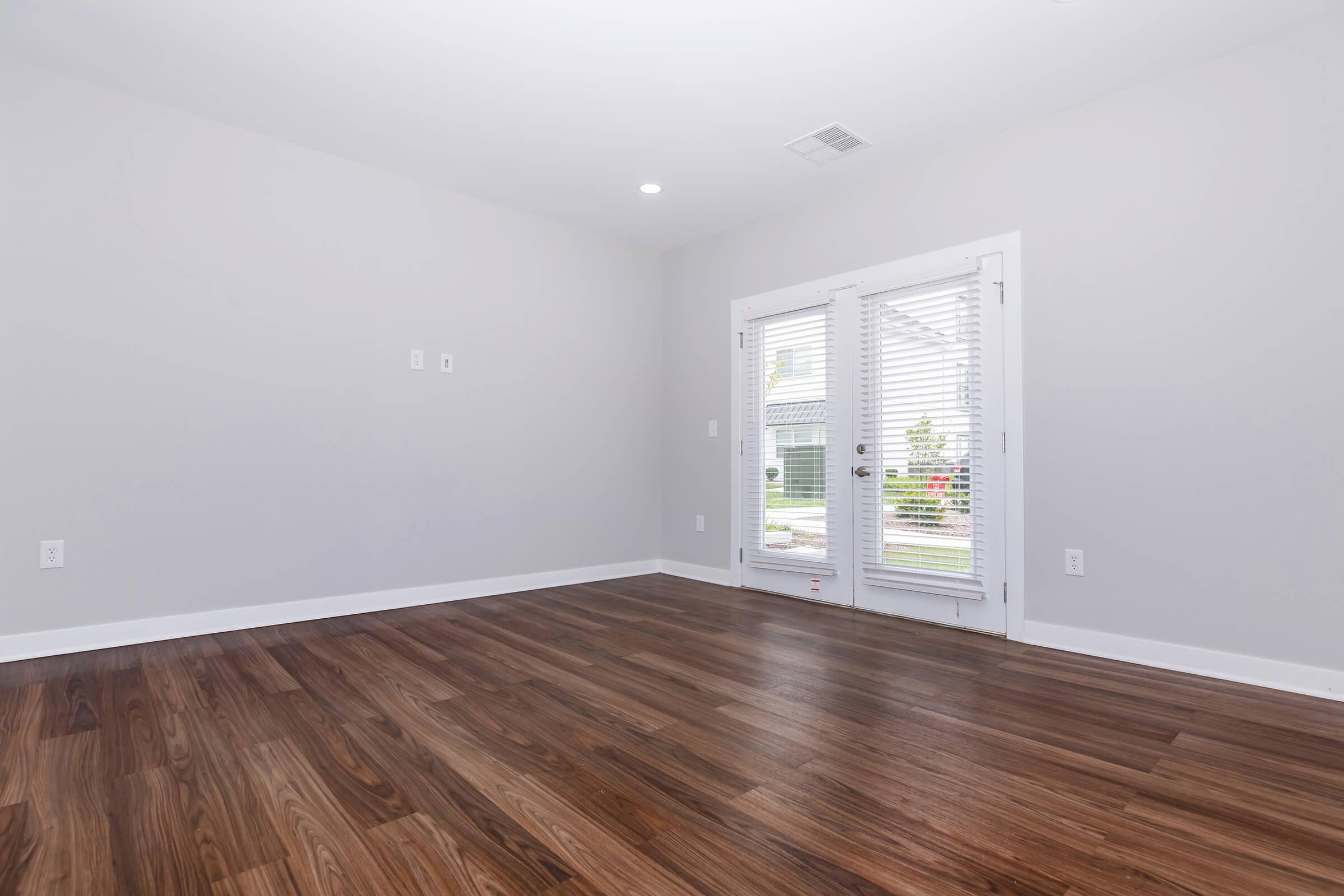 Empty room with light gray walls and a wooden floor. Two large glass doors with white blinds lead to an outdoor area, allowing natural light to enter. The space is modern and unfurnished, creating a clean, open atmosphere.