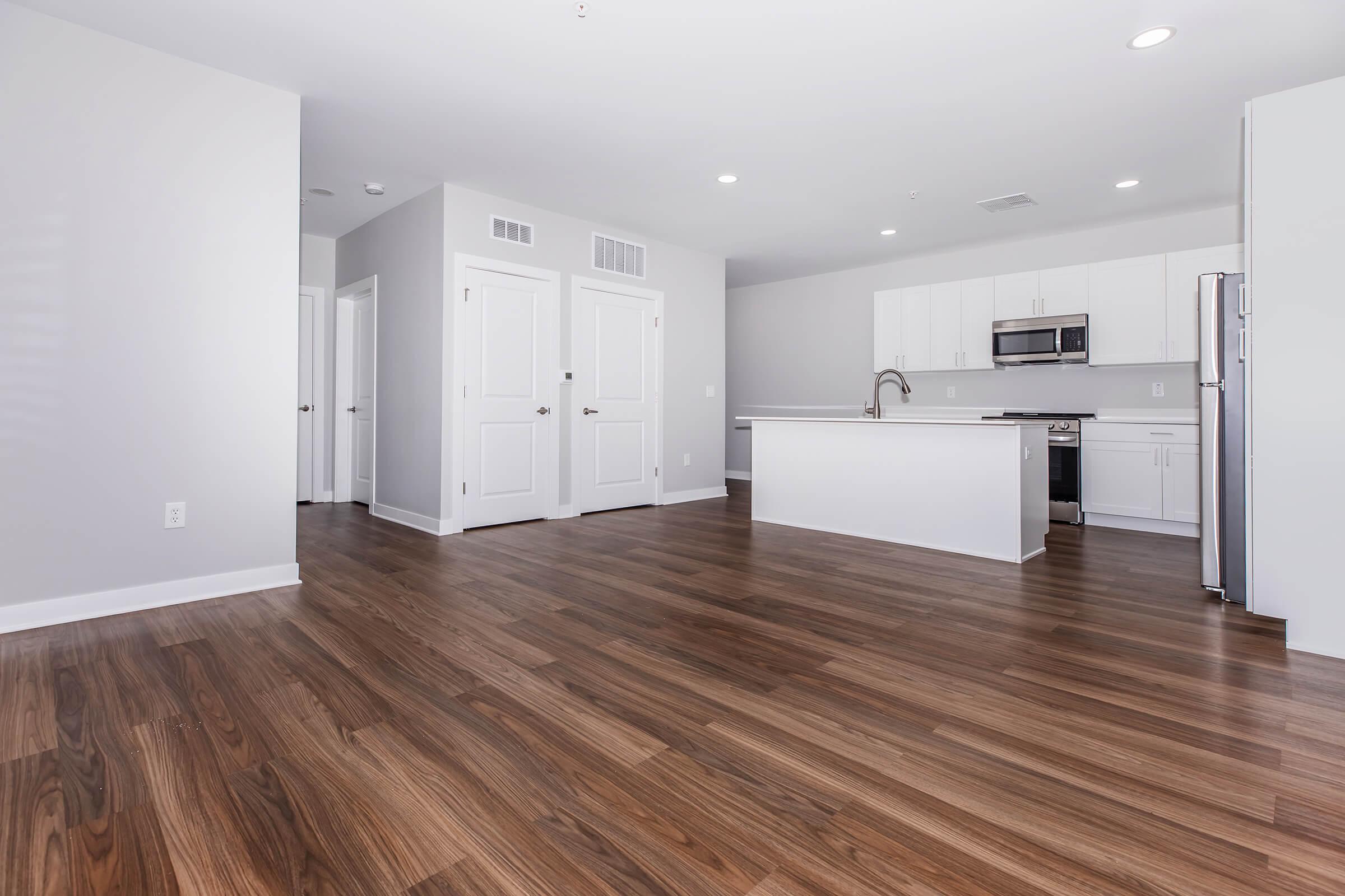 Spacious modern apartment interior featuring hardwood floors, white walls, and an open layout. The kitchen, equipped with stainless steel appliances and a large island, is visible in the background. Multiple doorways lead to other rooms, creating a bright and airy atmosphere.
