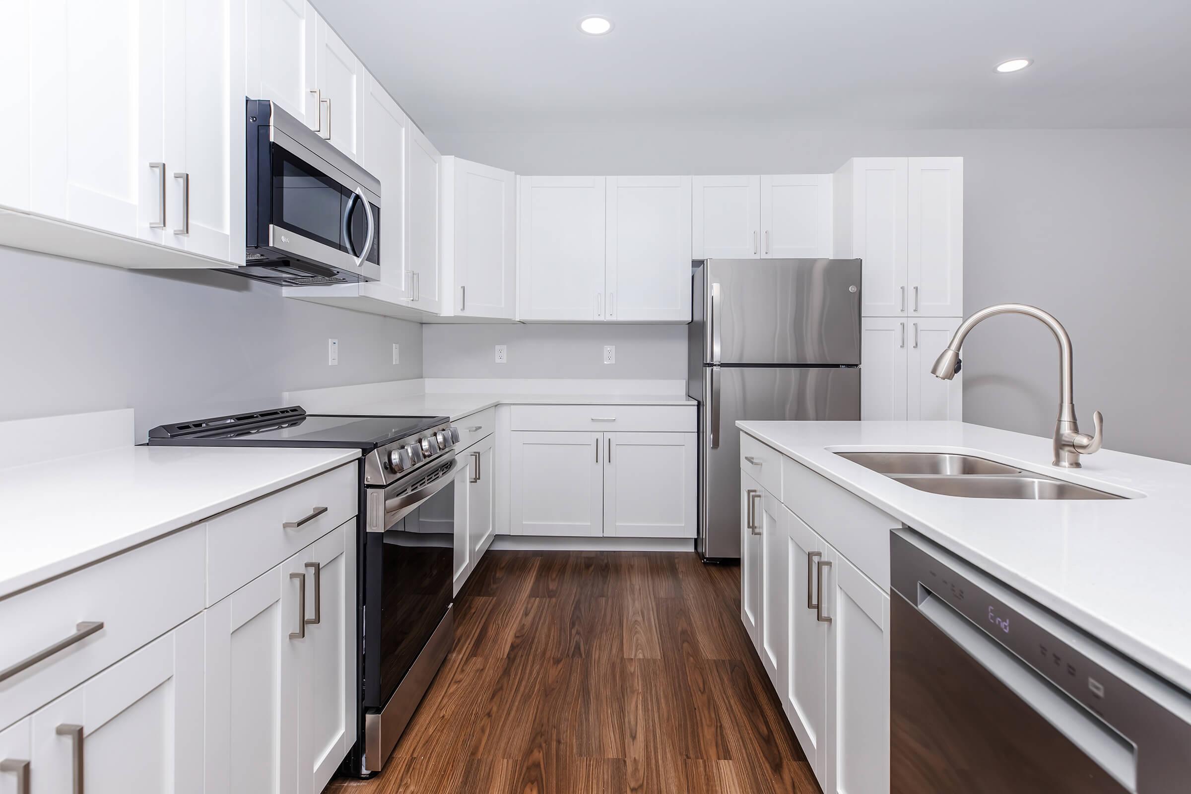 Modern kitchen featuring white cabinetry, stainless steel appliances including a microwave and refrigerator, a gas stove, a large sink with a sleek faucet, and hardwood flooring. The space is well-lit with recessed ceiling lights and has a contemporary design.