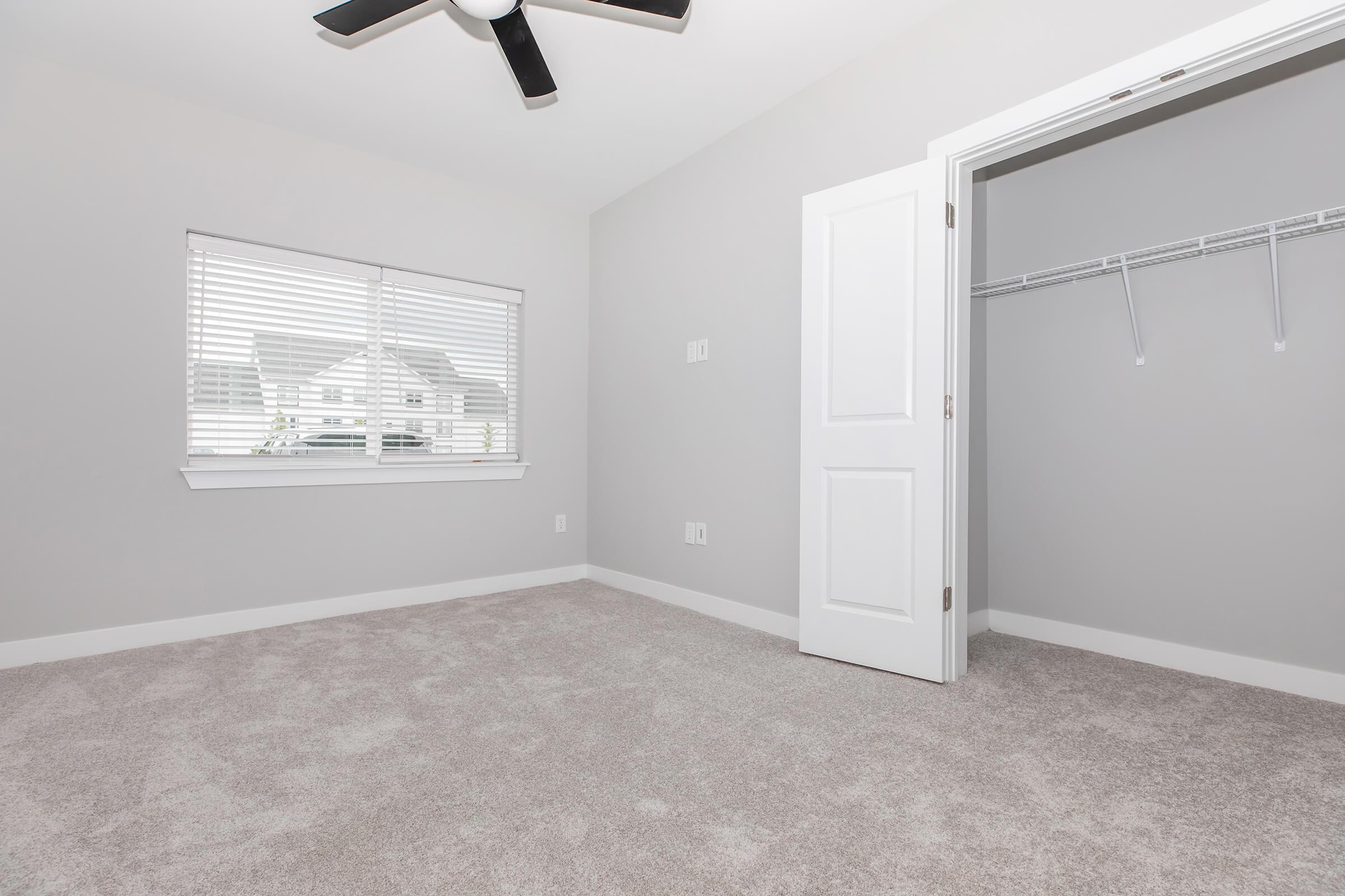 A well-lit empty room featuring light gray walls and carpeted flooring. There is a window with blinds allowing natural light in, and an open closet with a hanging rod. A ceiling fan is installed above, providing a modern touch to the space.