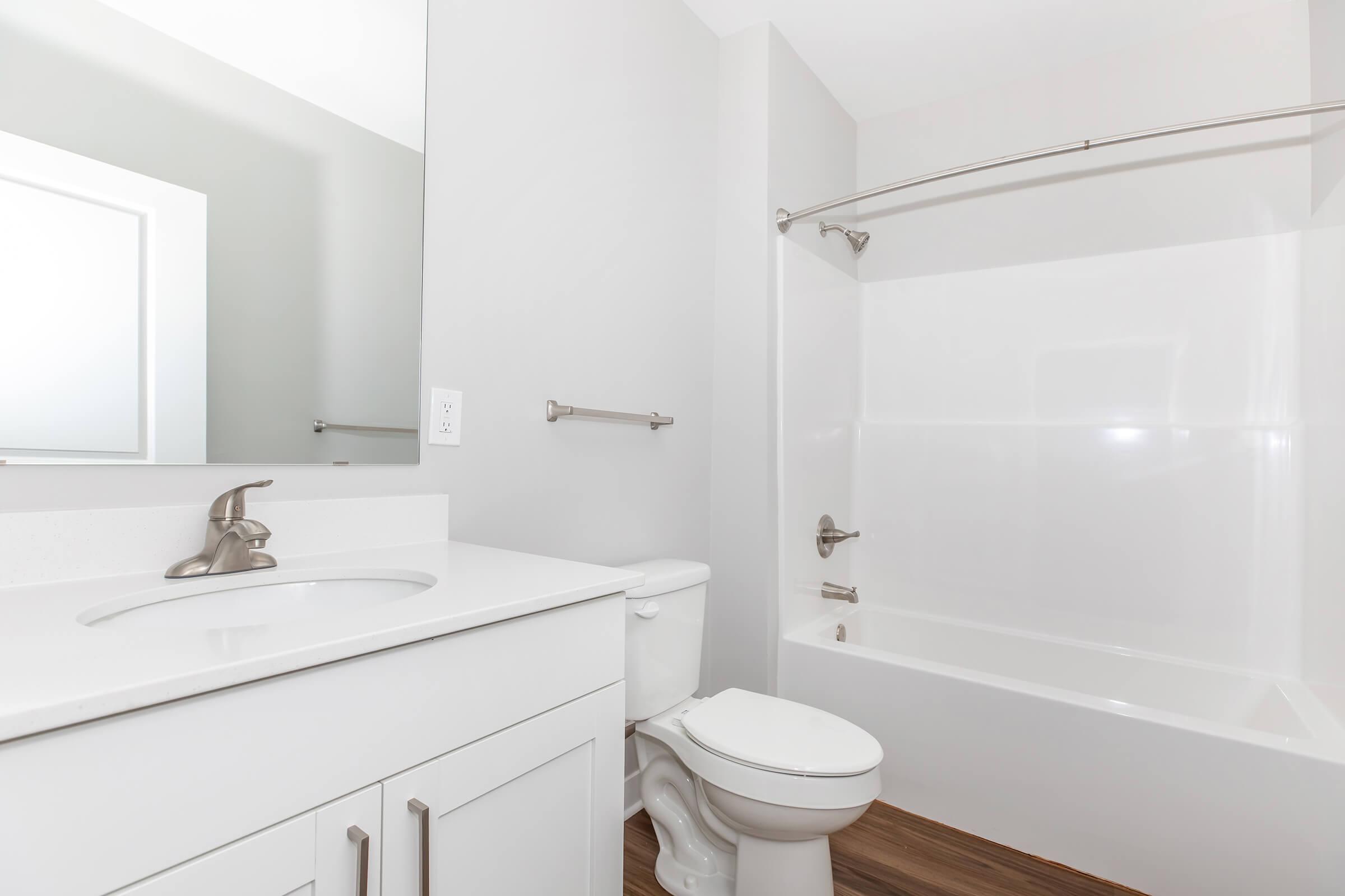 A clean and modern bathroom featuring a white bathtub with a shower curtain, a white vanity with a sink, and a large mirror. The walls are painted in a light gray color, and the flooring has a warm wood-like finish. The space is bright and well-lit, creating a fresh and inviting atmosphere.