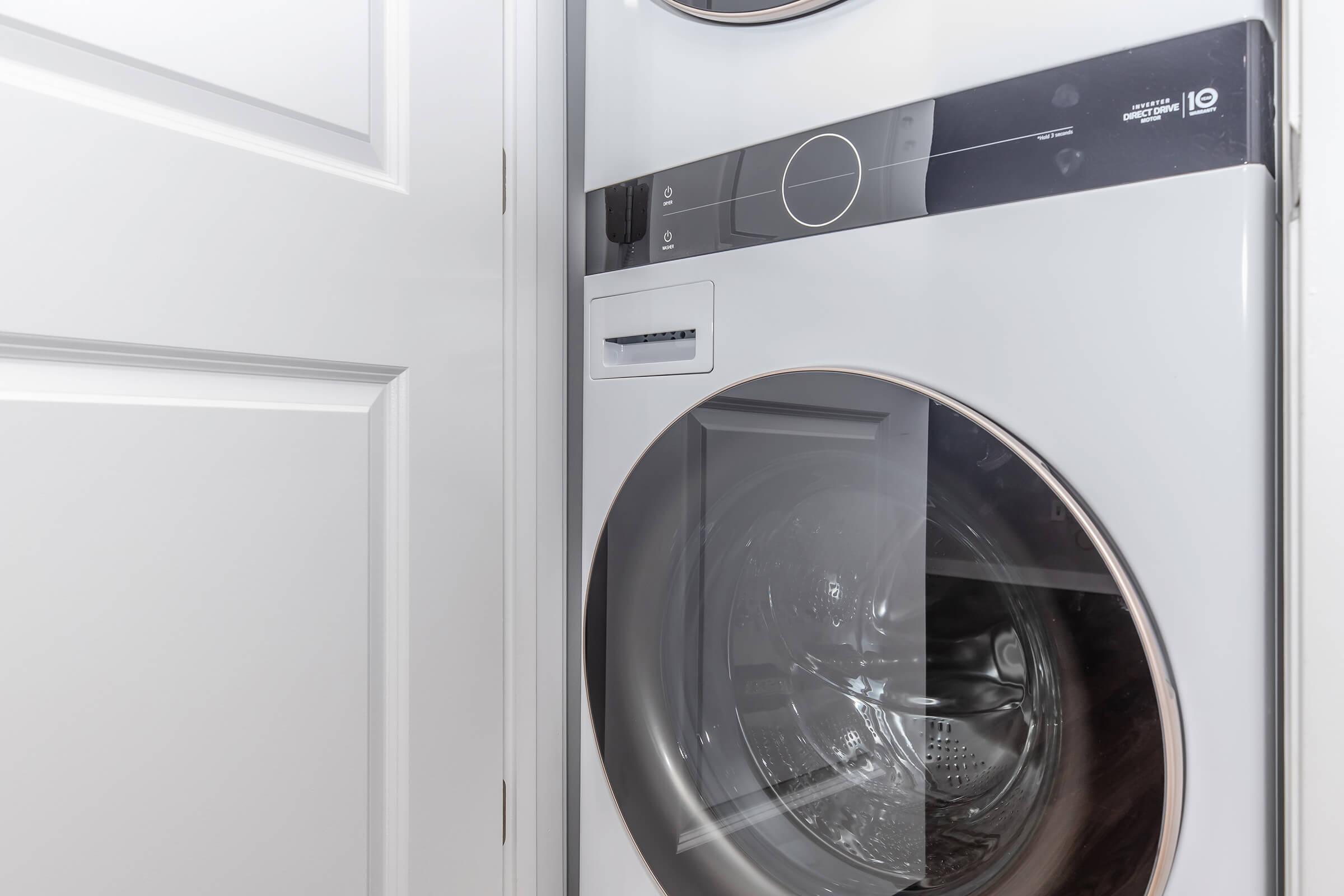 A modern, front-loading washing machine and dryer combo located in a laundry room, partially enclosed by white cabinet doors. The machines feature a sleek design with a digital control panel and a transparent door showcasing the interior drum.