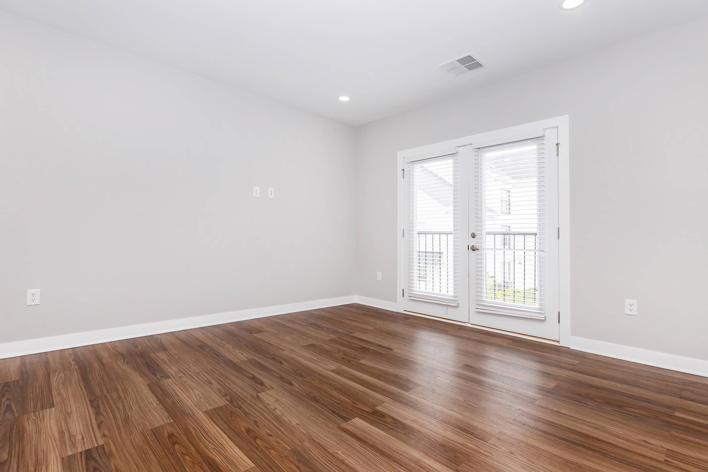 Empty interior room with light gray walls and wooden flooring. There are two French doors with white blinds leading to a balcony. The space is well-lit with recessed lighting in the ceiling.