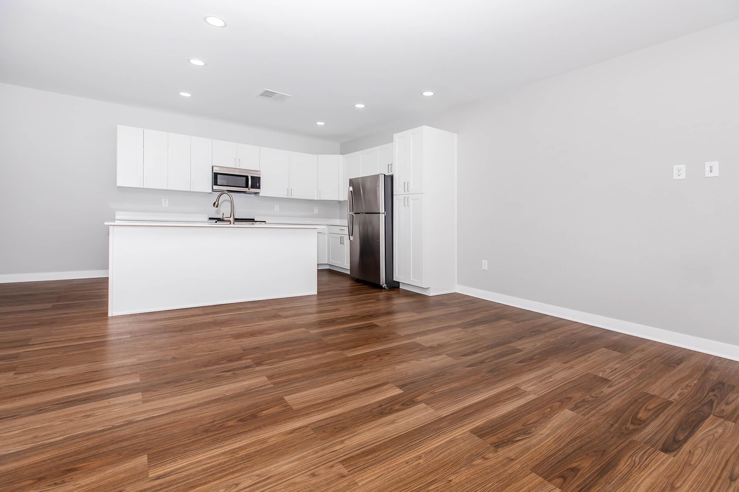 A modern, spacious kitchen and living area with light wood flooring. The kitchen features white cabinets, stainless steel appliances, and a central island with a sink. Bright, natural lighting shines through recessed lights, creating an inviting atmosphere. The walls are painted in neutral tones.