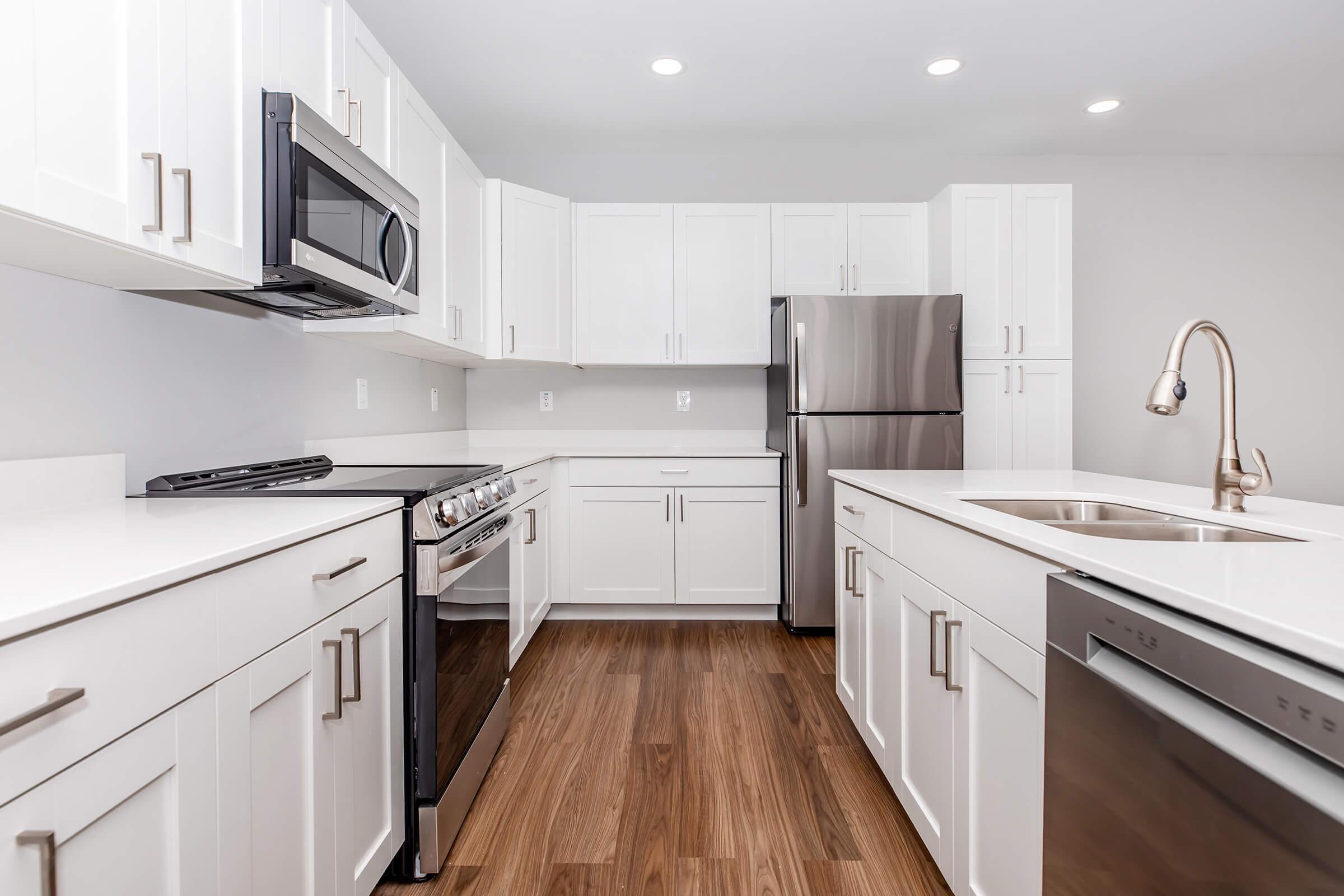 Modern kitchen featuring white cabinetry, stainless steel appliances, and a sleek countertop. Includes a gas range, microwave, refrigerator, and dishwasher. The space is well-lit with recessed lighting and has a warm wood floor, giving it a contemporary feel.