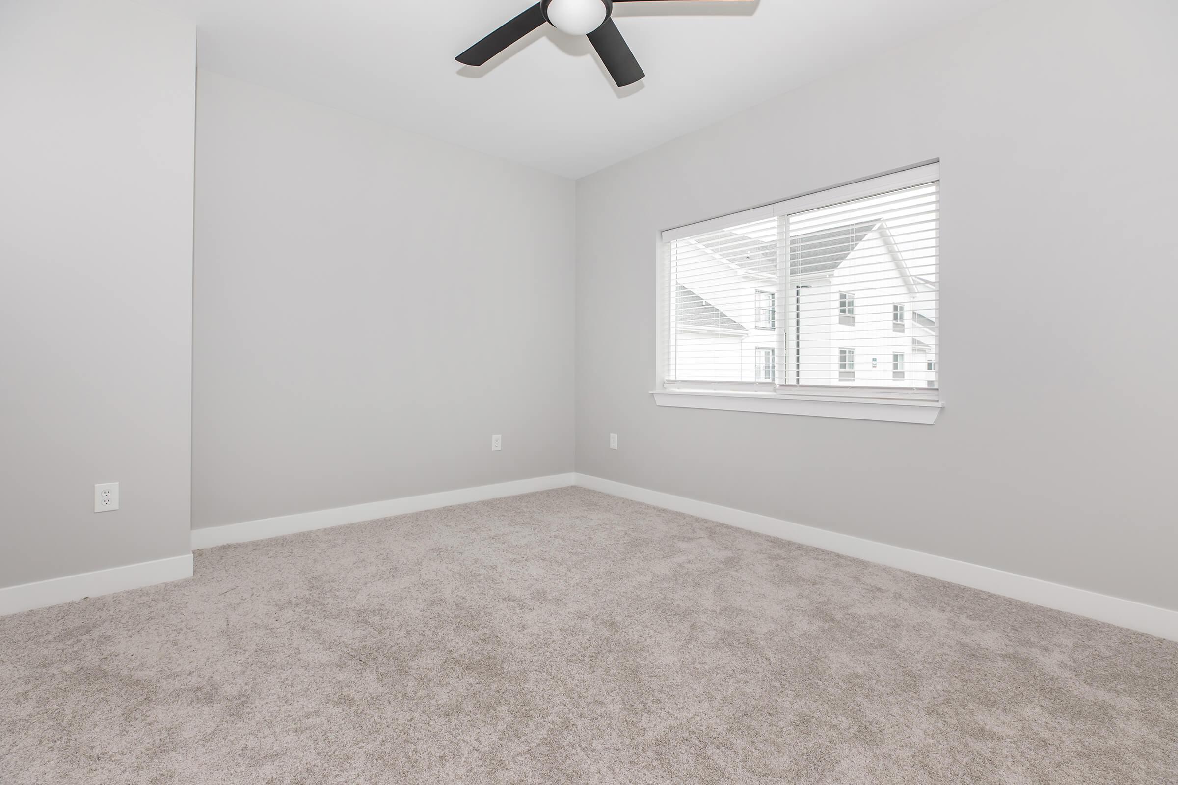 Empty room with gray walls and a light gray carpet. There's a ceiling fan with black blades and a window with white blinds allowing natural light to enter. The view outside shows the sides of adjacent houses.