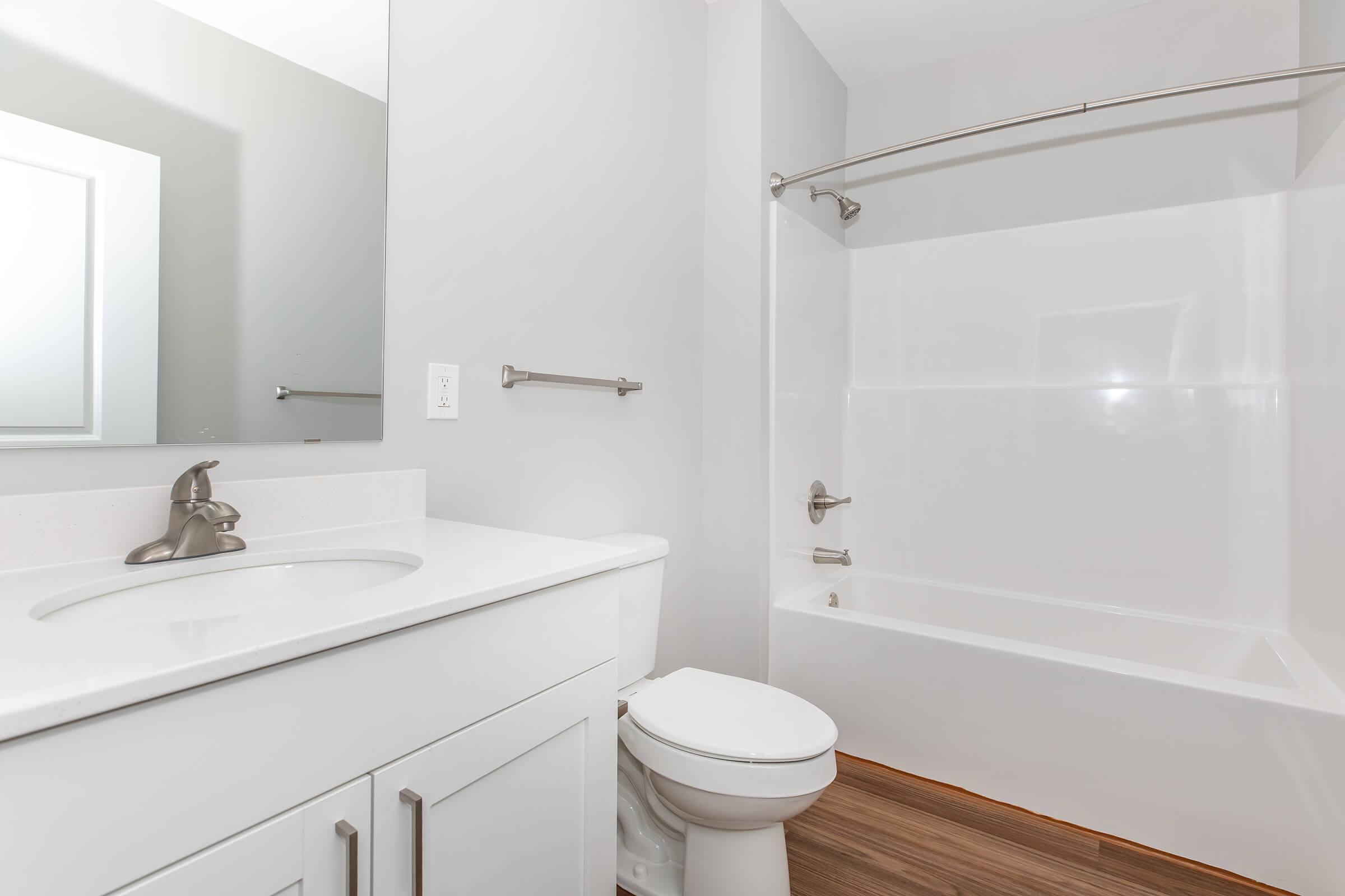 A modern bathroom featuring a white bathtub with a shower rod, a clean white countertop with a sink, a wall mirror, and light gray walls. The floor is covered with wooden planks, and there's a towel bar on the wall.