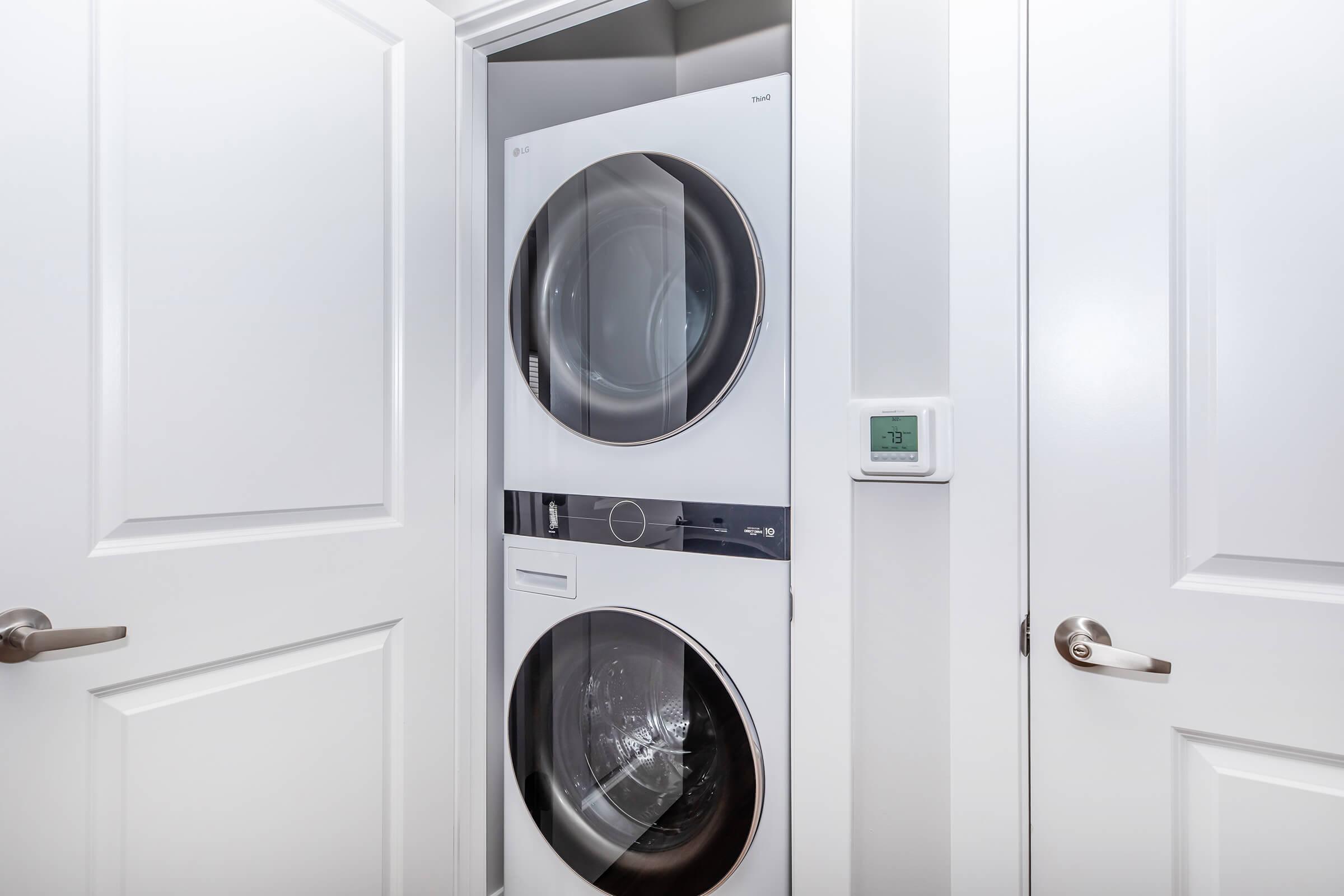 A modern laundry setup featuring a stacked washer and dryer in a white closet. The doors are closed, and a digital thermostat or control panel is visible on the wall beside the appliances. The overall design is sleek and contemporary, emphasizing a clean, organized space.