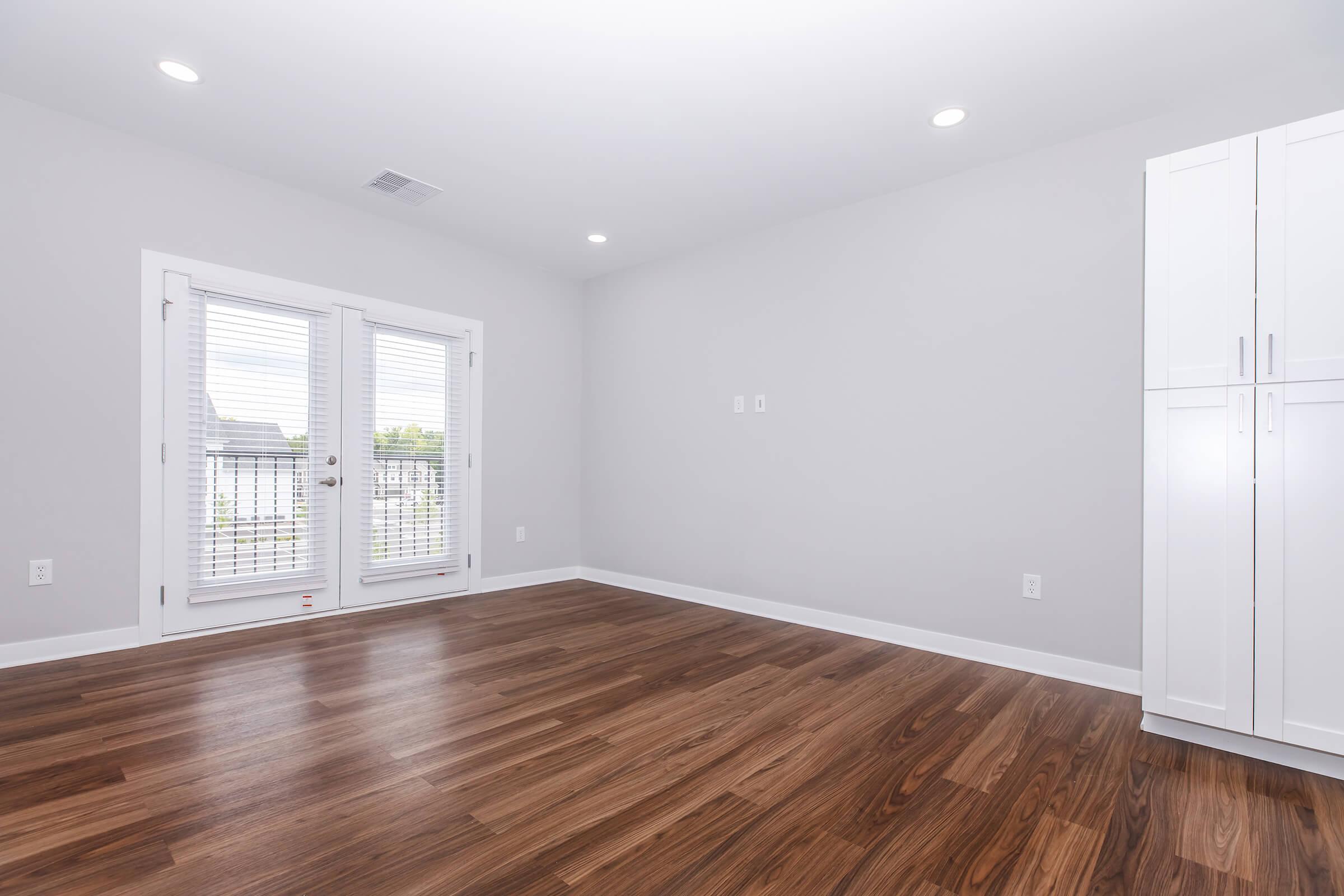 A bright, empty room with light gray walls and polished wooden flooring. Features large, sliding glass doors that lead to a balcony, framed by white molding. On one side, there is a tall white cabinet against the wall, providing storage space. Ample overhead lighting creates an open and airy atmosphere.