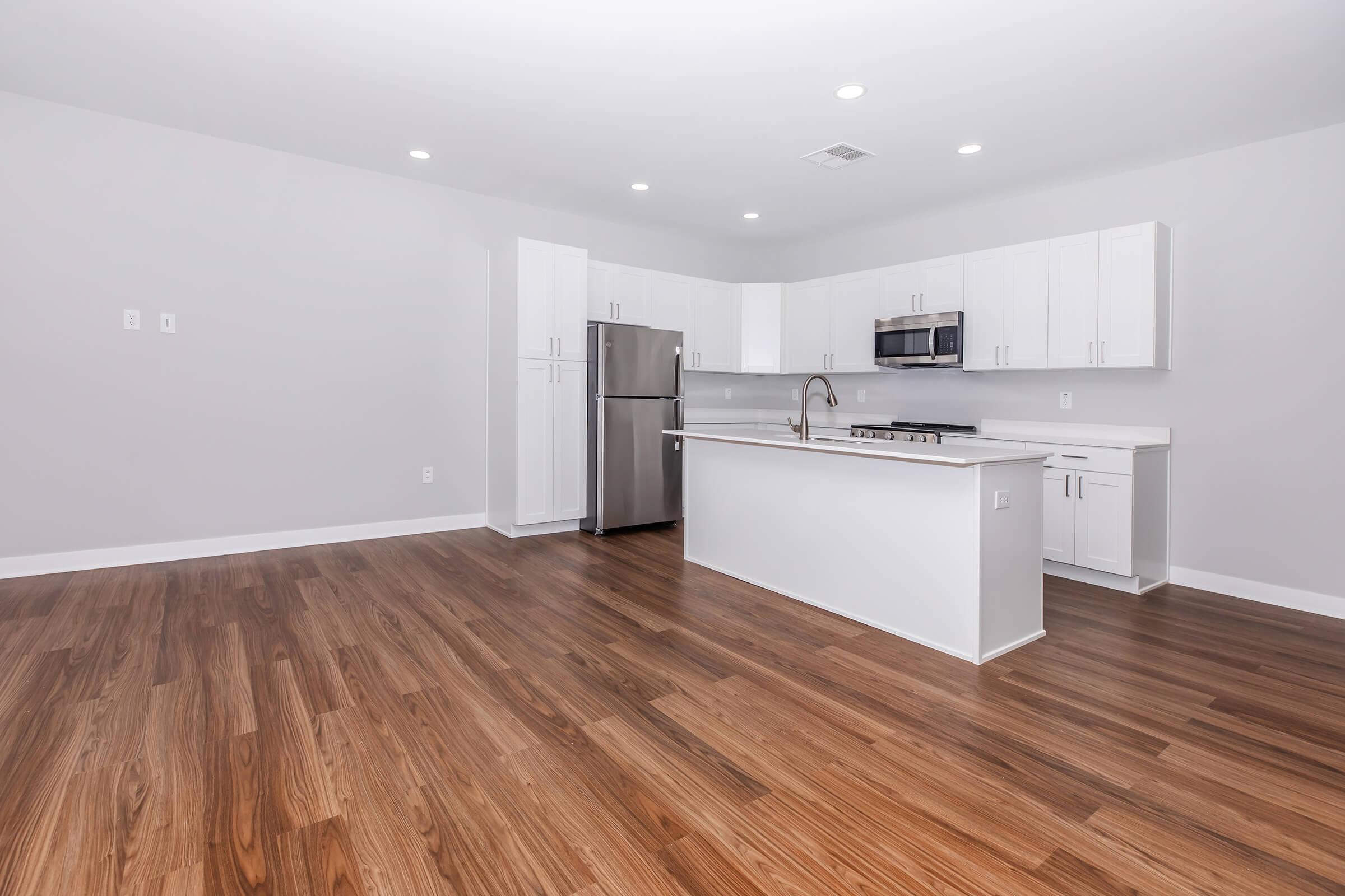 A modern kitchen and living area with light-colored walls, sleek white cabinetry, stainless steel appliances, and a wooden floor. The kitchen features an island with a sink and a stovetop, while the spacious living area is empty, creating an open and airy look. Bright overhead lighting complements the design.
