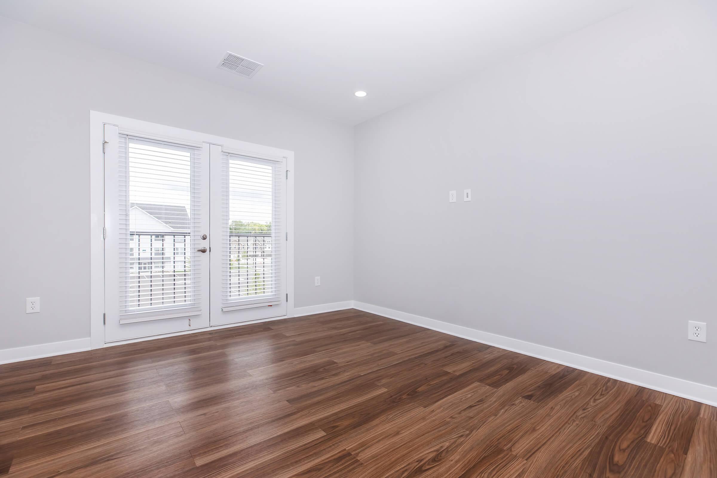Empty room with light gray walls and a wood flooring. There are large double doors with blinds that lead to a balcony or outside area. The ceiling has recessed lighting, and there are two small wall outlets visible. The space is bright and uncluttered, giving a modern and fresh appearance.