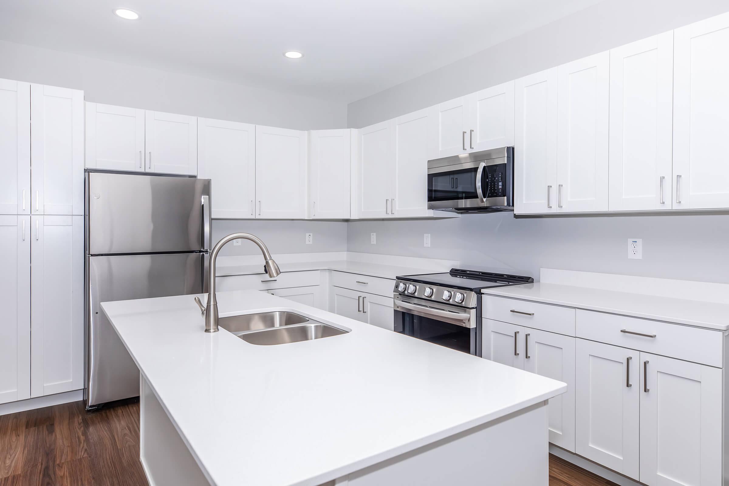 Modern kitchen featuring white cabinetry, stainless steel appliances including a refrigerator and microwave, and a gas range with an oven. The kitchen island has a built-in sink, with a clean, uncluttered design and hardwood flooring. Natural light fills the space from overhead lighting.
