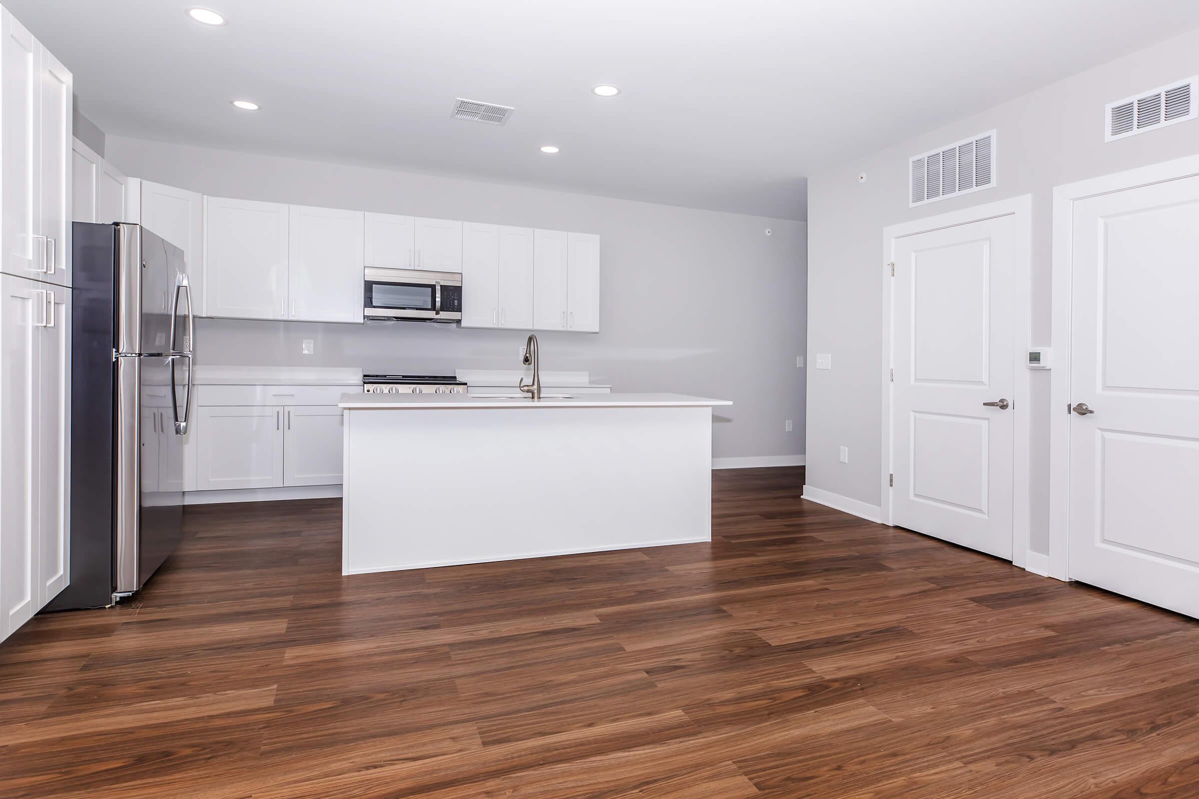 Modern kitchen with white cabinets, stainless steel appliances, and a large island. The space features hardwood flooring and neutral-colored walls, illuminated by overhead lighting. The open layout includes a door leading to another room and a sleek design suitable for contemporary living.