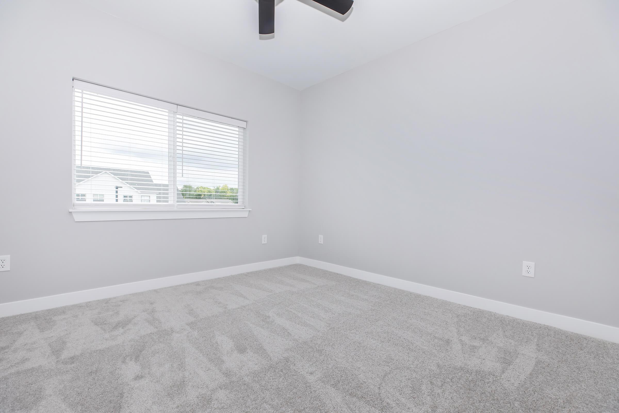 A clean, empty room with light gray walls and carpeting. There is a window with white blinds on one wall, allowing natural light to enter. The ceiling features a black ceiling fan, and the overall ambiance is spacious and ready for personalization.