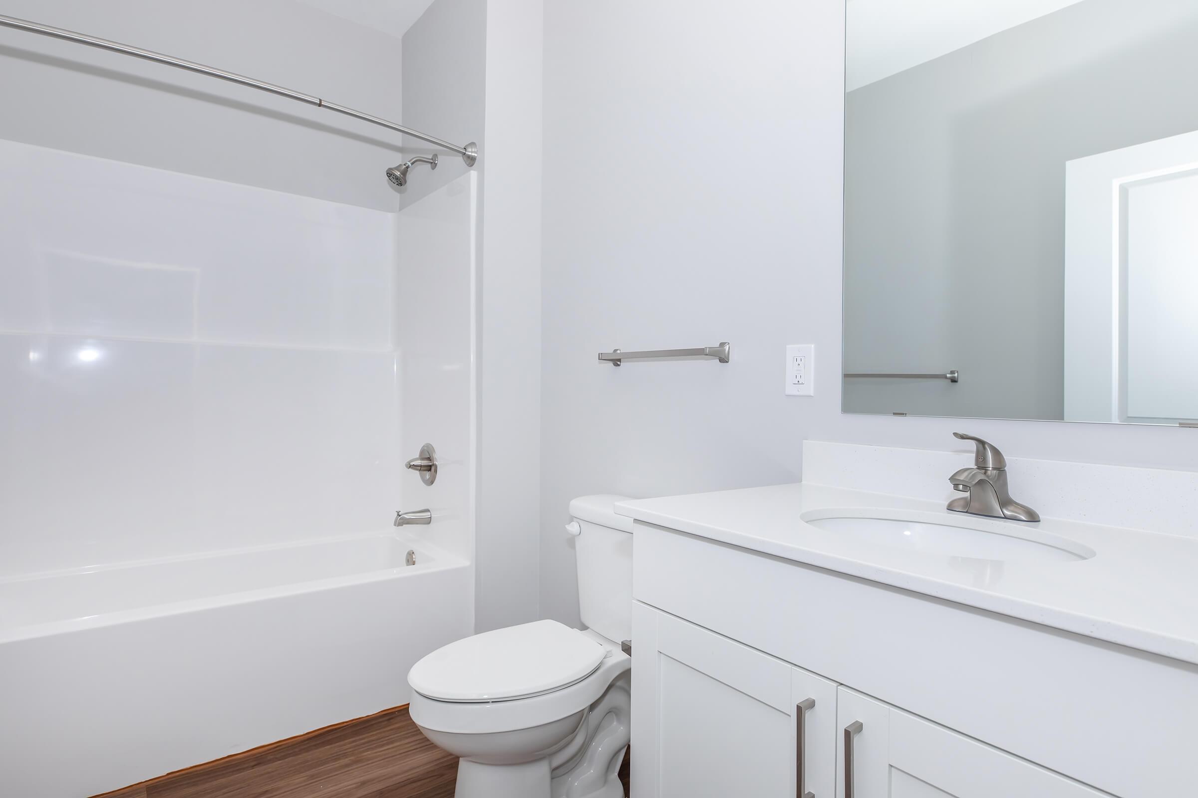 A clean and modern bathroom featuring a white bathtub with a silver shower rod, a white toilet, and a vanity with a sink. The walls are painted light gray, and a large mirror is mounted above the sink. The floor has a wood-like appearance, adding warmth to the space.