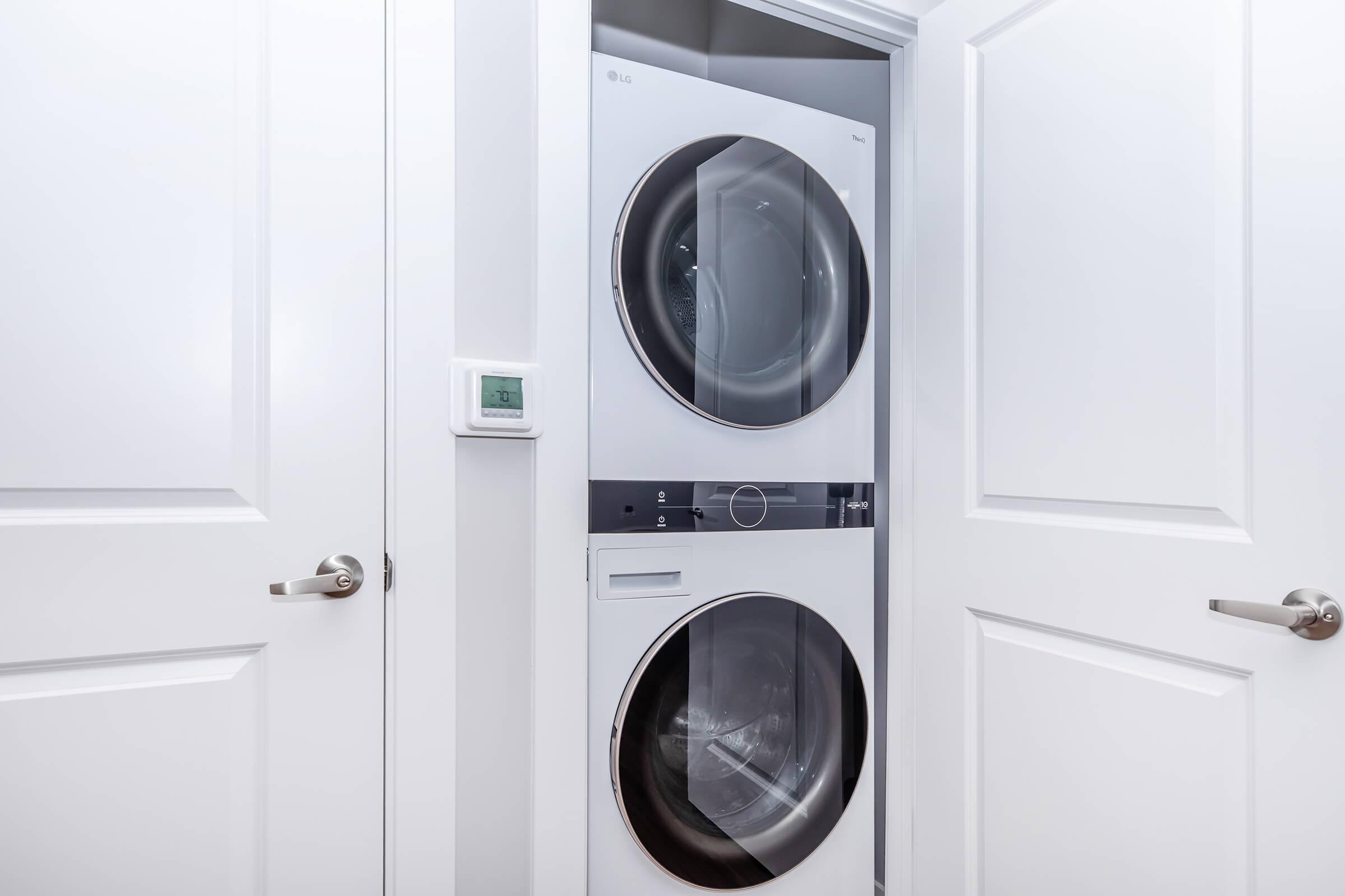 A modern laundry room featuring a stacked washer and dryer from LG, housed in a white closet with double doors. The interior is well-lit, highlighting the sleek design of the appliances, with a thermostat visible on the wall beside the units.