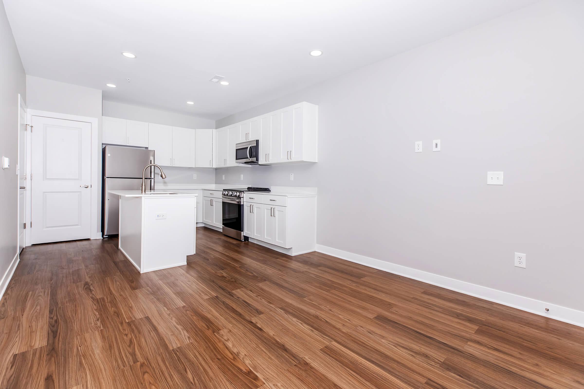 A modern kitchen featuring white cabinetry, stainless steel appliances, and a central island. The space has light wood flooring and is brightly lit with natural light. The walls are painted a soft gray, providing a clean and contemporary aesthetic.