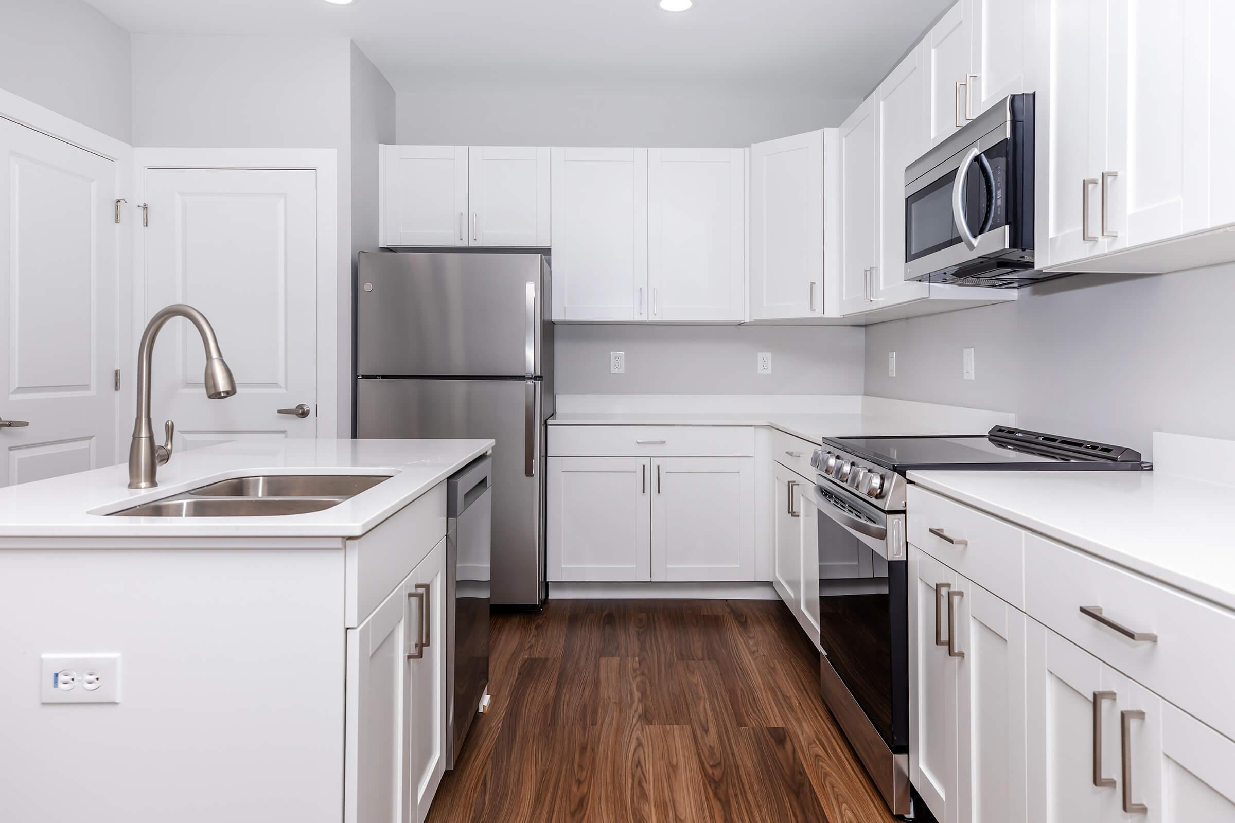 Modern kitchen featuring white cabinetry, stainless steel appliances including a refrigerator and oven, a double sink with a brushed nickel faucet, and a sleek design. The floor is made of wood, and the space is well-lit with recessed lighting.