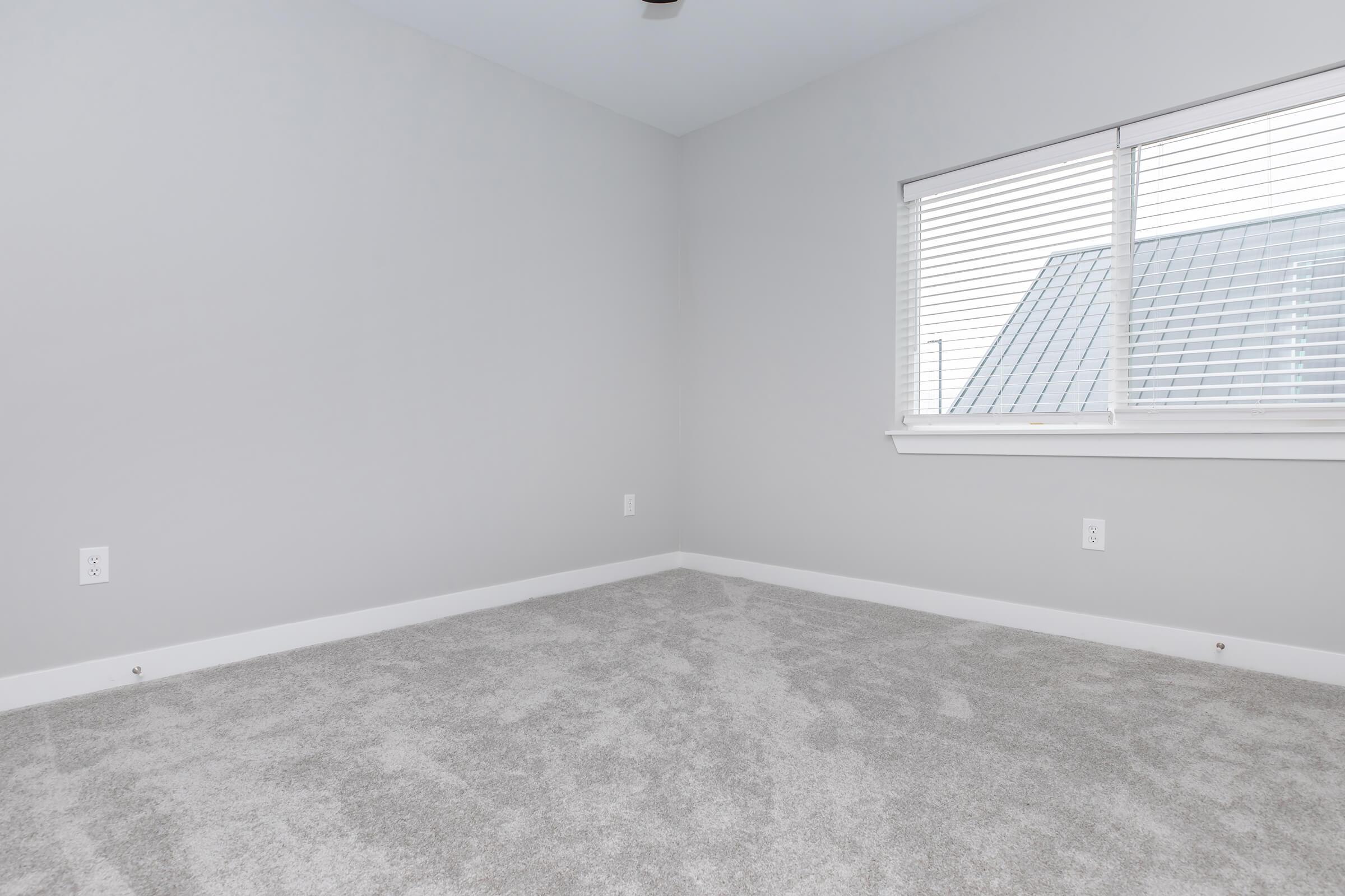 A spacious, empty room with gray walls and light gray carpet. There is a window with blinds on one wall, allowing natural light to enter. The room is unfurnished, creating a minimalist and clean appearance, suitable for various uses or decoration.