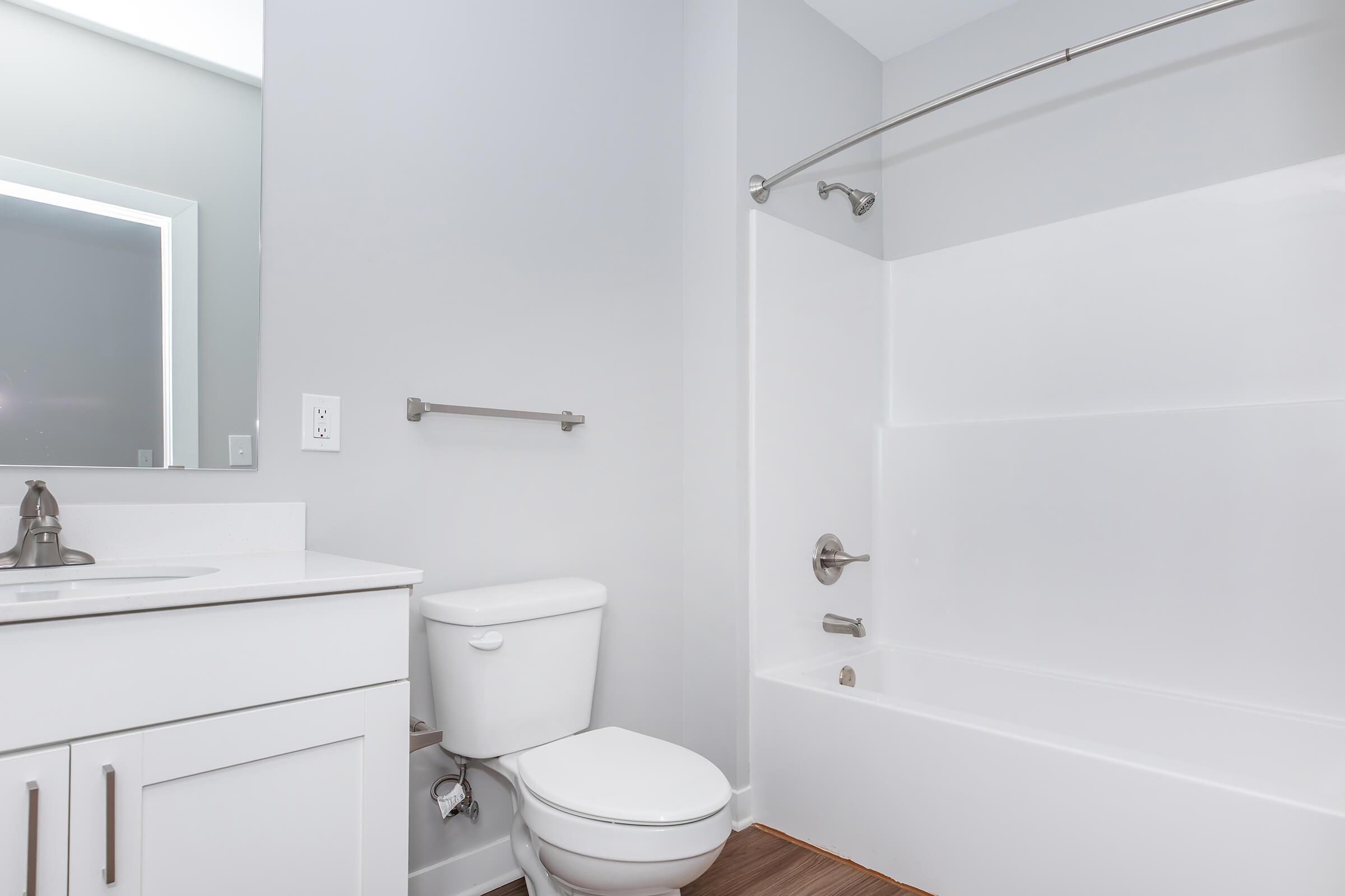 A clean, modern bathroom featuring a white bathtub and shower, a white toilet, and a vanity with a sleek sink. The walls are painted light gray, and there is a towel bar mounted on the wall. A large mirror reflects light, enhancing the bright and airy atmosphere of the space.
