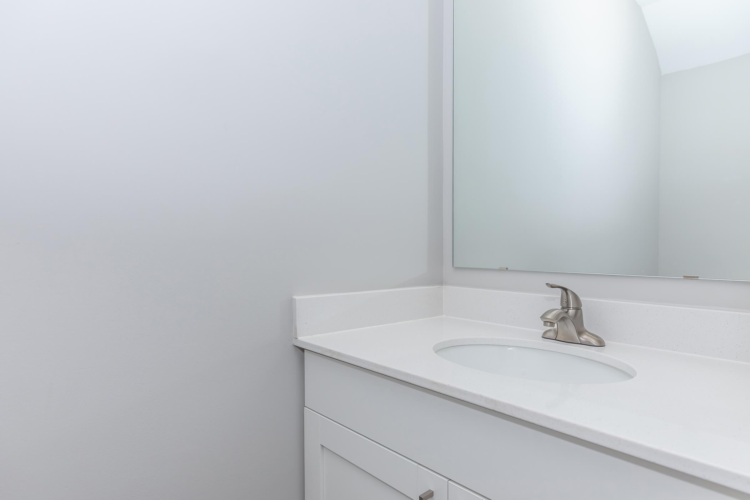 A clean and minimalistic bathroom sink area featuring a white countertop, a modern faucet, and a mirror. The walls are painted in a light color, creating a bright and airy atmosphere.
