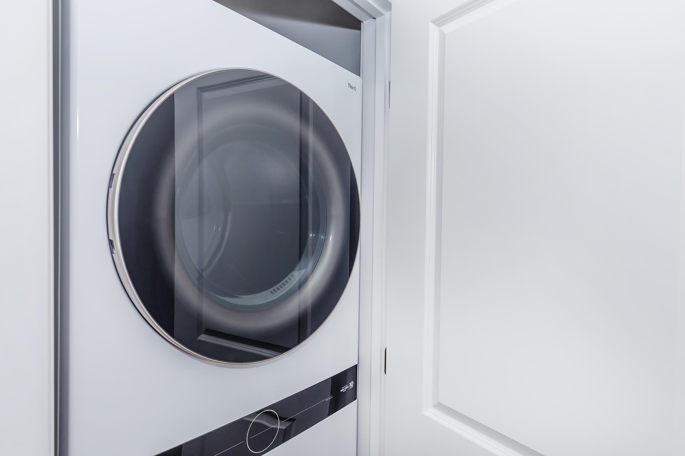 A modern dryer with a round glass door, located in a laundry room. The dryer has a sleek design with a dark front and a control panel at the bottom. A white door is partially open, providing a view into the laundry area.