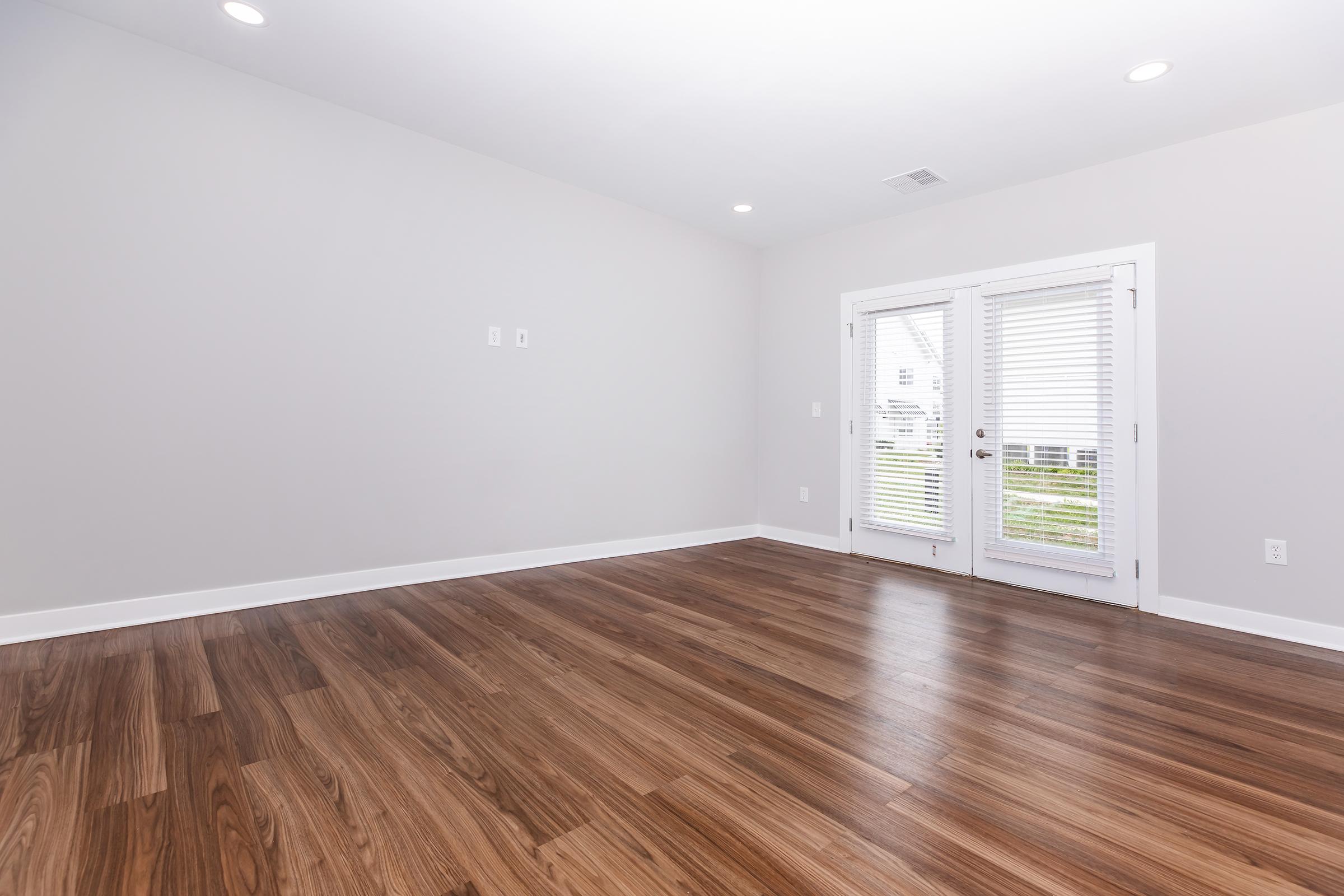 Empty interior room with light gray walls and beautiful hardwood flooring. A pair of glass French doors with white blinds opens to an outdoor area, allowing natural light to brighten the space. The room is spacious and unfurnished, giving a clean and modern feel.