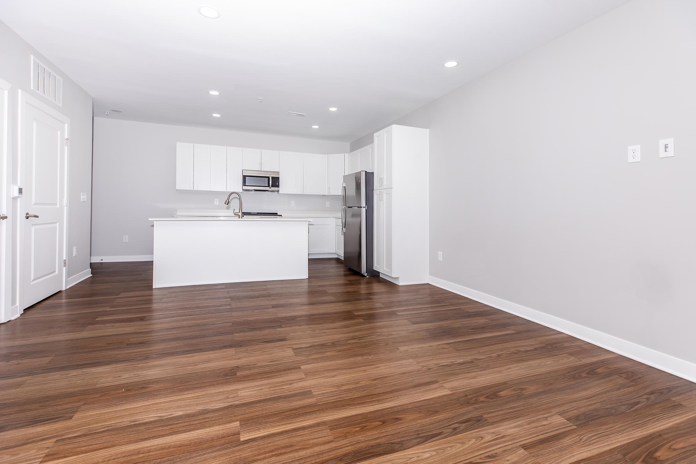 Modern kitchen and living area featuring light gray walls, hardwood flooring, and an open layout. The kitchen includes white cabinetry, stainless steel appliances, a large island, and recessed lighting. The space is well-lit with natural light and has no furniture, creating a spacious feel.