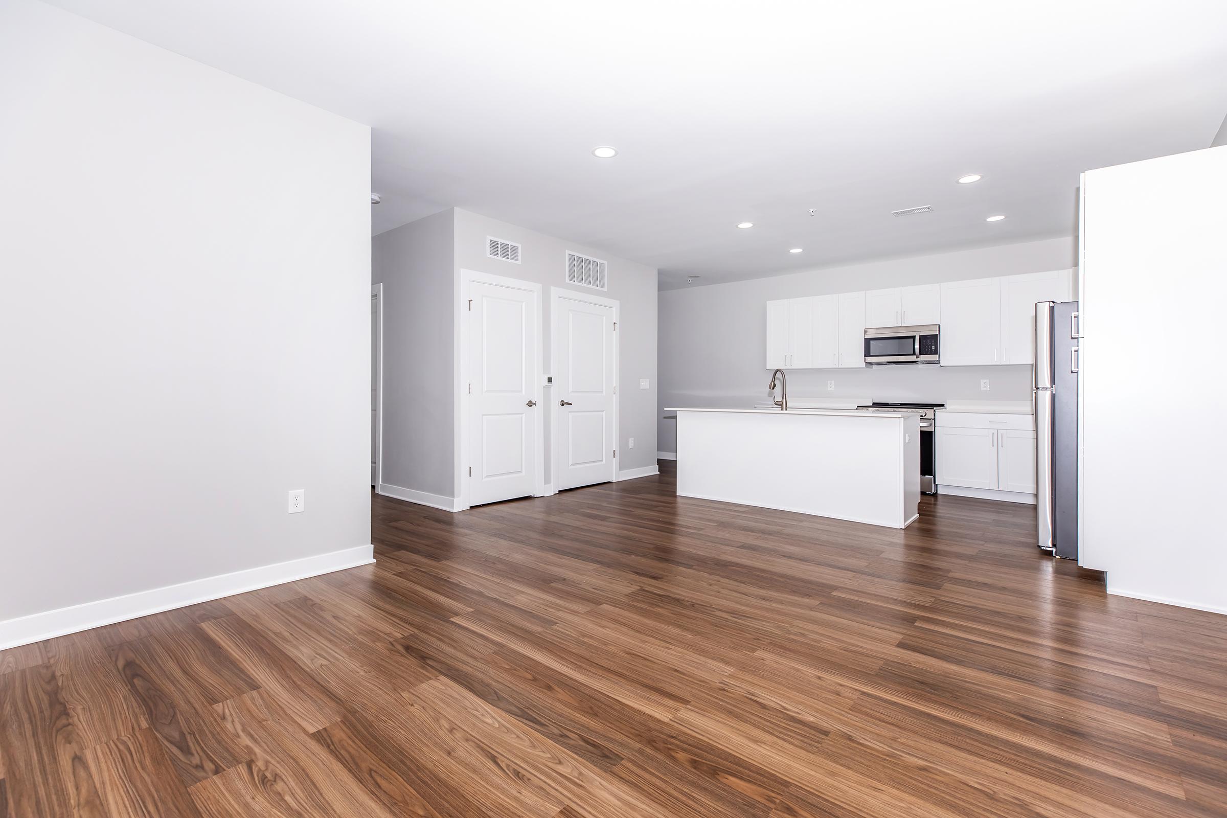 A bright, modern apartment interior featuring hardwood flooring, white walls, and a spacious open layout. The kitchen includes stainless steel appliances and a large island. The room is well-lit with multiple lights and has two closed doors leading to other areas.
