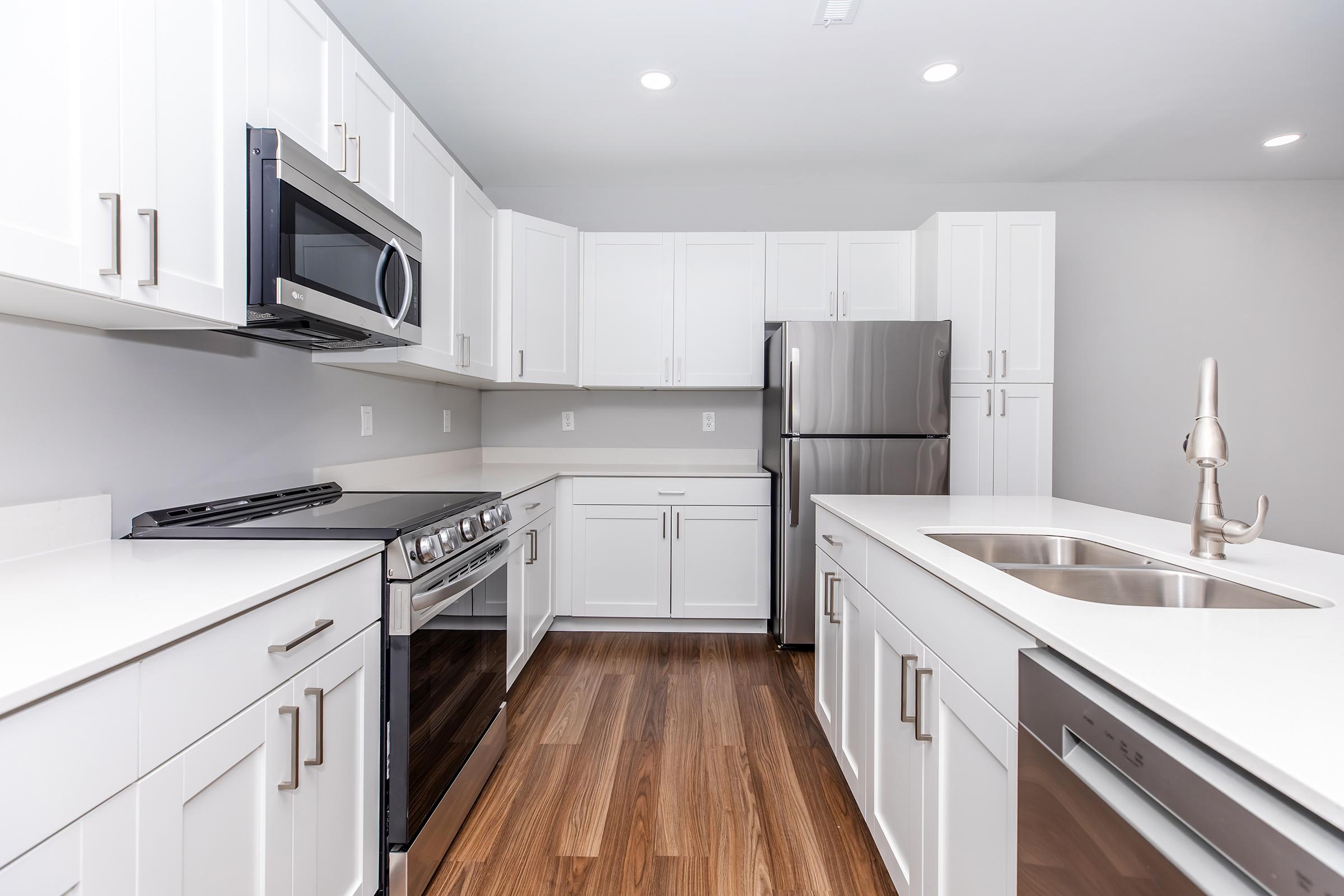 Modern kitchen featuring white cabinetry, stainless steel appliances including a microwave, oven, and refrigerator, along with a double sink. The countertops are light-colored, and the flooring is wood, creating a bright and spacious atmosphere. Recessed lighting enhances the contemporary design.
