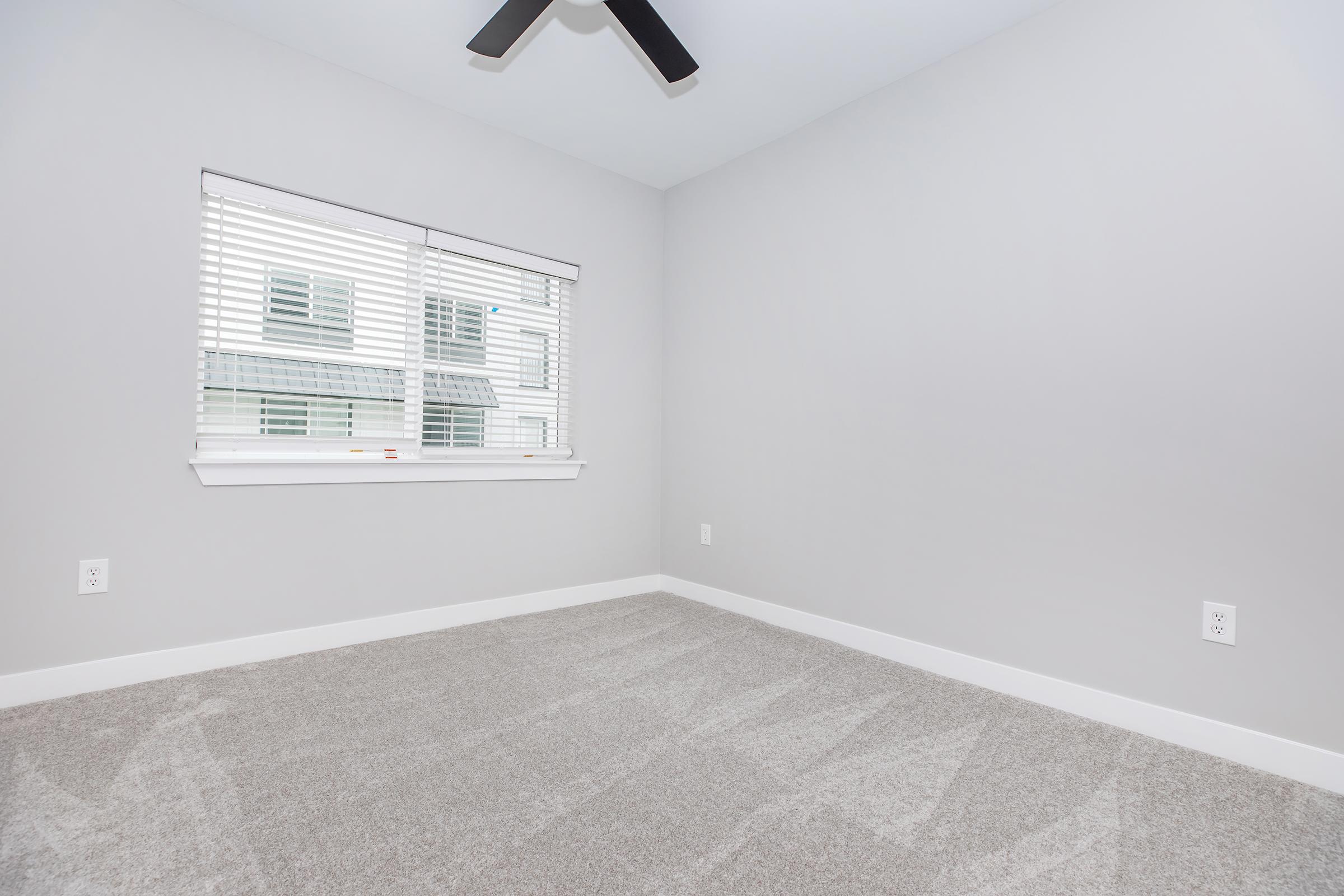 A light gray empty room with a ceiling fan, featuring white walls and a window with blinds. The floor is covered in soft gray carpet, and there are no furnishings or decor present. Natural light filters through the window, illuminating the space.